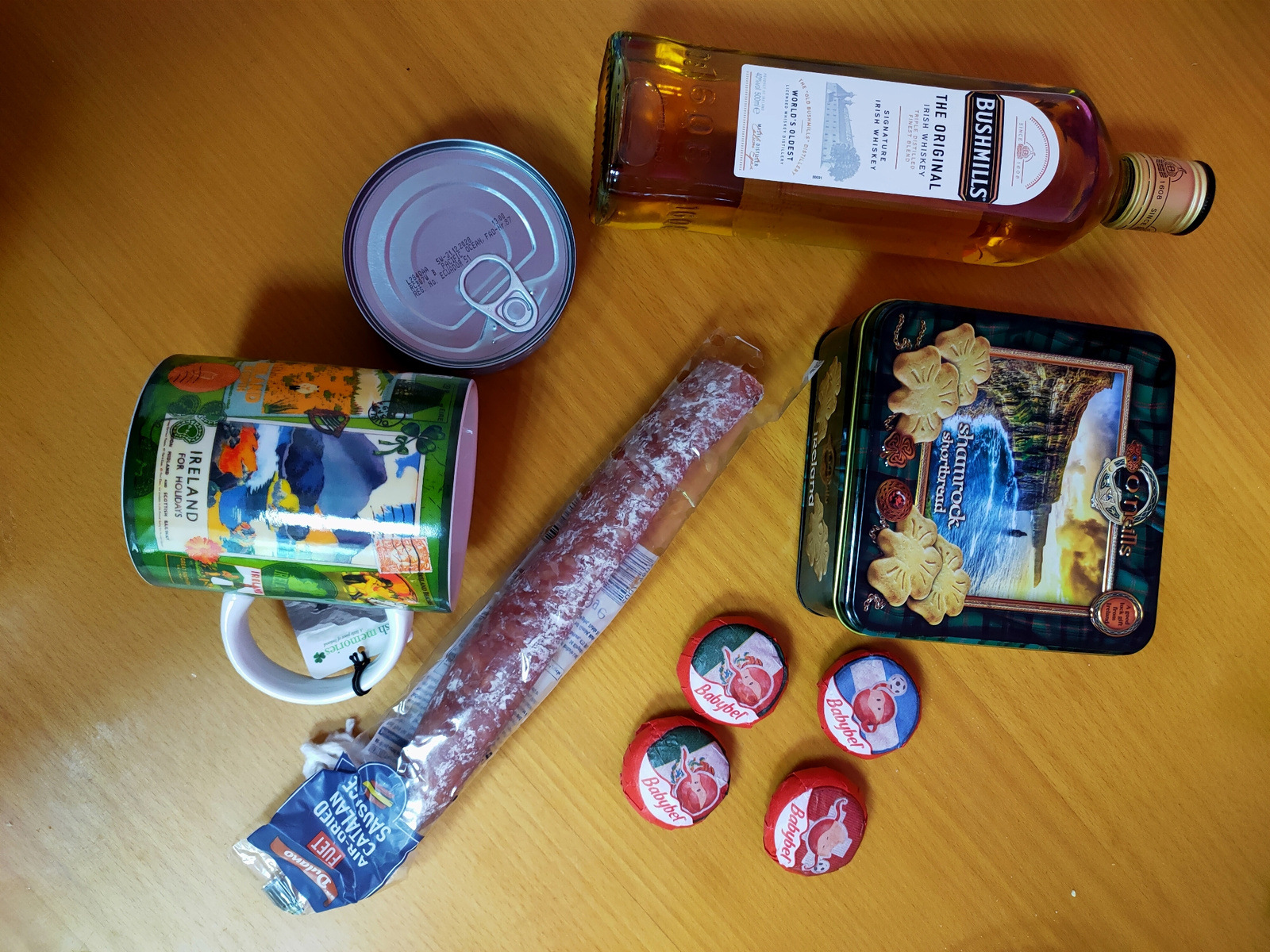 The image shows a collection of food and drink items laid out on a wooden surface. The items include a bottle of Bushmills Irish Whiskey, a can, a tin of shortbread, a packaged air-dried Catalan sausage, and four Babybel cheese portions. A white mug with an Irish design is also present. The scene is lit from above, casting shadows on the surface. The overall impression is that of a curated collection of goods, possibly for a gift or a meal. The image does not provide any information about the city of Kremenchuk, Ukraine, or any specific location.