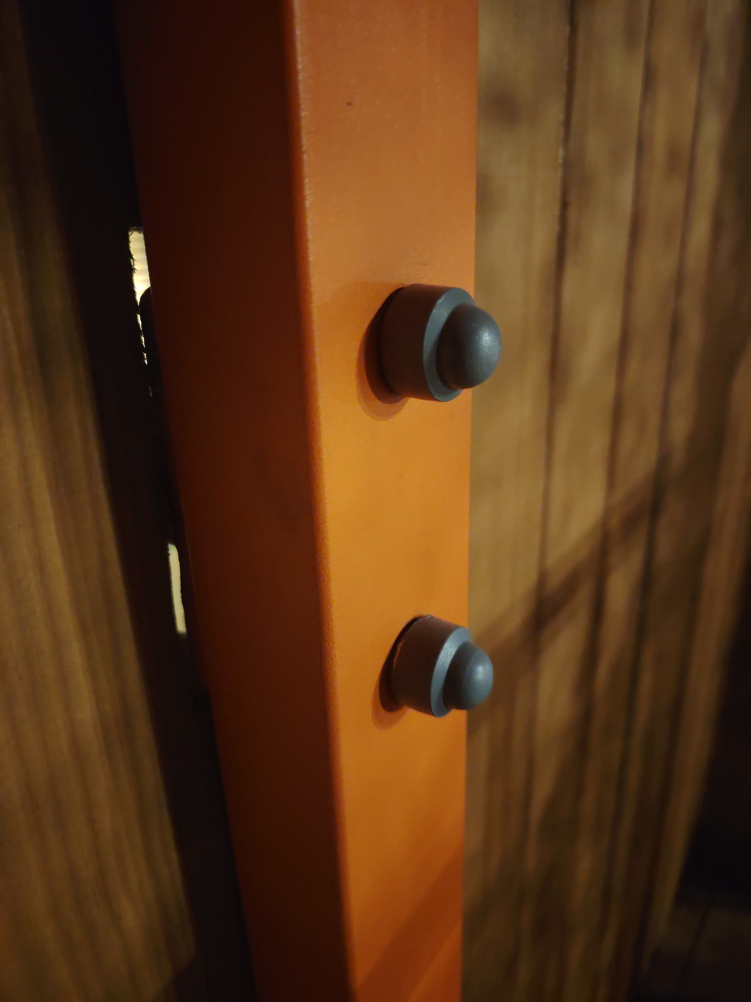 The image is a close-up shot of an orange, rectangular post with two grey, rounded knobs or buttons attached to its side. The knobs are positioned vertically, one above the other. The post is angled, and the background is a blurred, wooden surface with vertical grain patterns. The lighting creates shadows on the wooden background. There are no people visible in the image. The image does not contain any specific geographical markers to determine its location within Yekaterinburg, Russia.