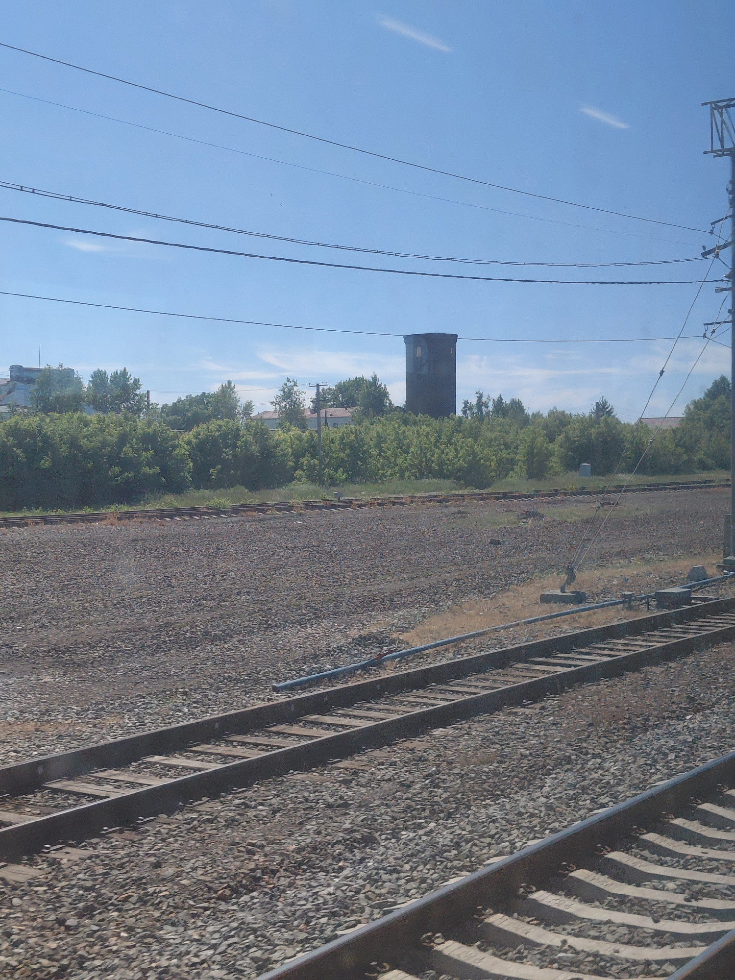 This image depicts a railway scene with several tracks running parallel to each other. The foreground is dominated by the ballast of the railway, with wooden sleepers visible beneath the metal rails. In the middle ground, there is a dense line of green trees, behind which are some buildings, including what appears to be a tall, cylindrical structure resembling a water tower. The sky is a clear blue with a few faint wispy clouds. Overhead, power lines are strung across the frame. The scene suggests an industrial or semi-rural area, likely on the outskirts of a settlement. Based on the provided context, this scene is located in the city of Chulym, Russia. There are no people or animals visible in the image.