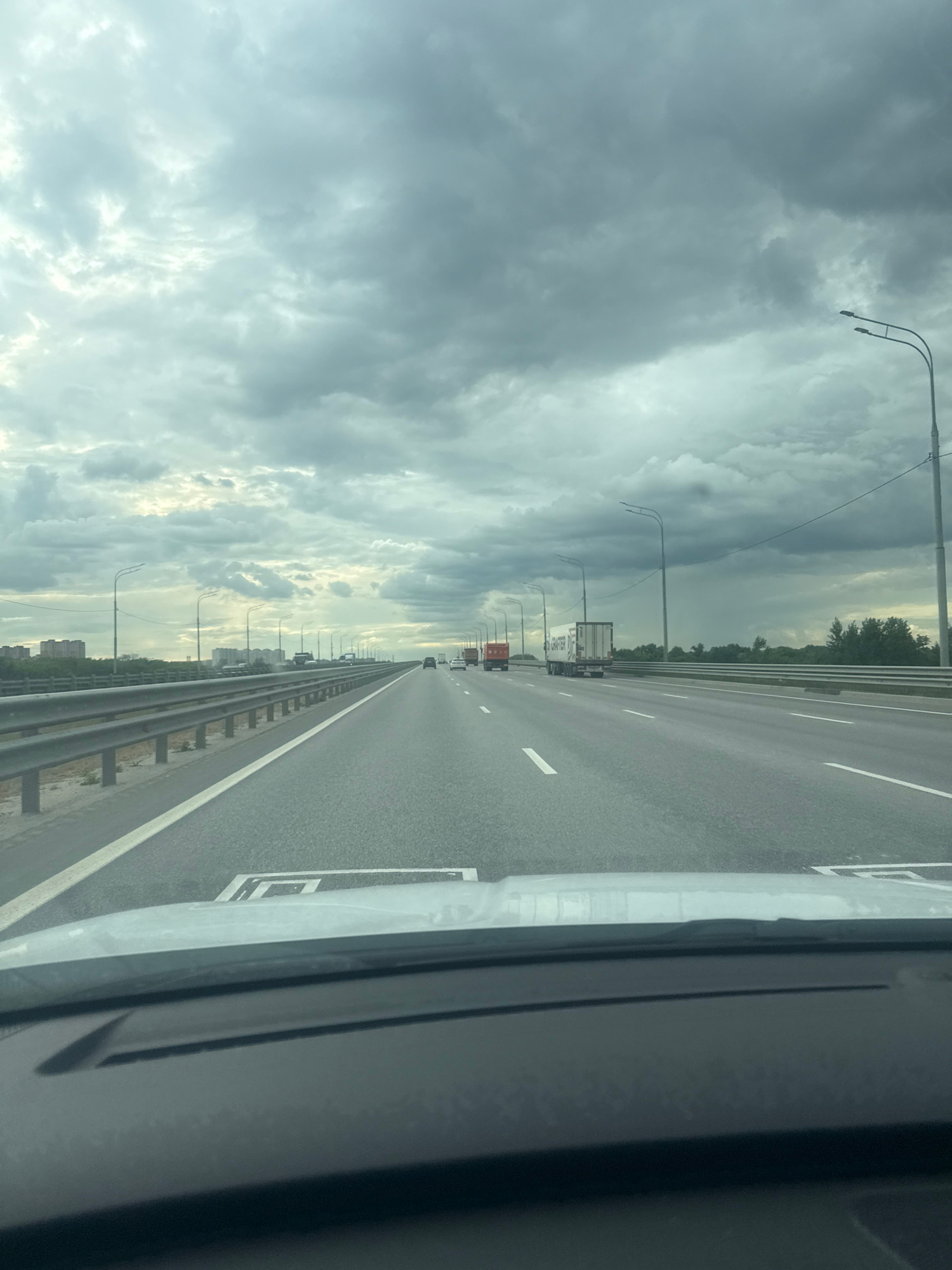 The image captures a view from inside a vehicle driving on a multi-lane highway under a cloudy sky. The highway appears to be an elevated road or a bridge, with metal guardrails lining the right side. Several vehicles, including trucks and cars, are visible on the road ahead and in the distance. On the left side of the highway, there are buildings and trees. The sky is filled with dark, heavy clouds, suggesting impending rain or overcast weather. Streetlights are positioned along the highway. The foreground shows a portion of the vehicle's dashboard and windshield. The scene is likely a highway outside of a city.