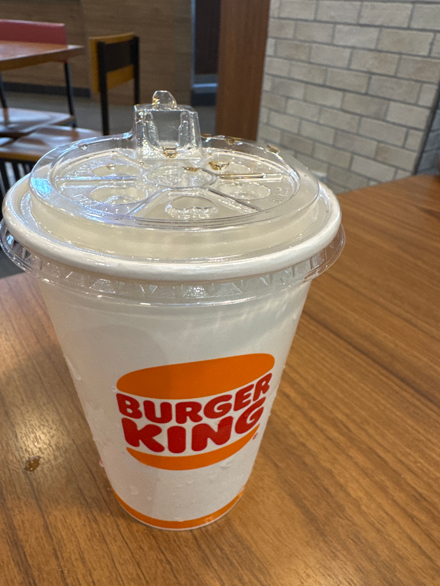 A close-up shot features a Burger King branded cup filled with a light-colored beverage, possibly lemonade or soda, with condensation visible on its exterior, suggesting it is chilled. The cup has a clear plastic lid with a raised section for a straw. The cup rests on a wooden table, on which a few small droplets of liquid are present. In the background, a blurred view of a restaurant interior includes tables, chairs, and a brick wall, consistent with a Burger King outlet. The lighting suggests an indoor setting during the day. There are no people or animals present in the image.