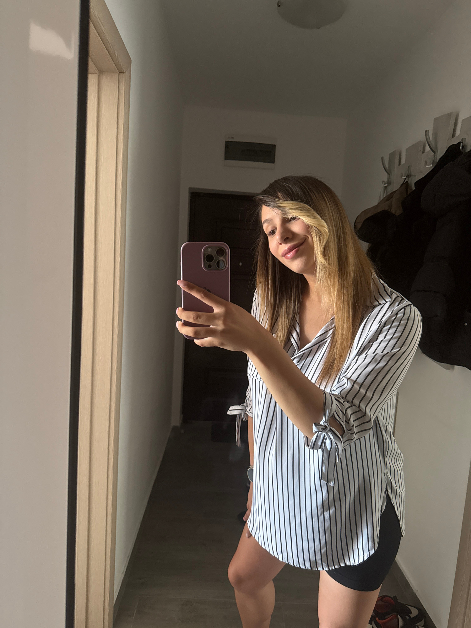 A young woman with blonde-streaked brown hair takes a selfie in a hallway with wooden doors and a large mirror. She is wearing a white and black pinstripe button-down shirt with tied sleeves and black shorts. She is holding a purple smartphone in her right hand. A coat rack with several jackets is visible on the wall behind her. The scene appears to be an interior residential hallway.
