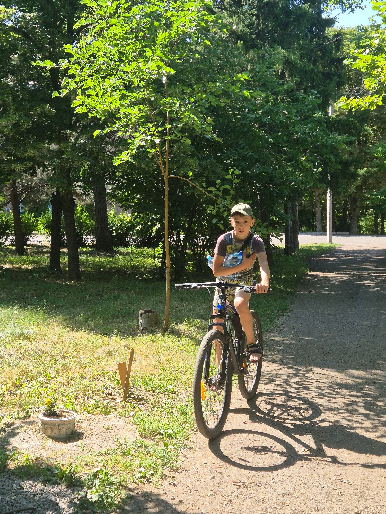A young boy, approximately 10-12 years old, rides a bicycle on a paved path in a park-like setting. He is wearing a green baseball cap, a t-shirt with a colorful, abstract pattern, and shorts. He has a plastic water bottle held in his left arm, which is slung across his chest. The bicycle is dark colored with black tires and silver rims. The path is dirt and gravel, shaded by trees on the left and right. Sunlight filters through the leaves, casting dappled shadows on the ground. The background consists of dense green trees. To the left of the path, there is a patch of dry, yellow grass with a small potted plant. The scene appears to be in the village of Sozonivka, Ukraine, on a sunny day.