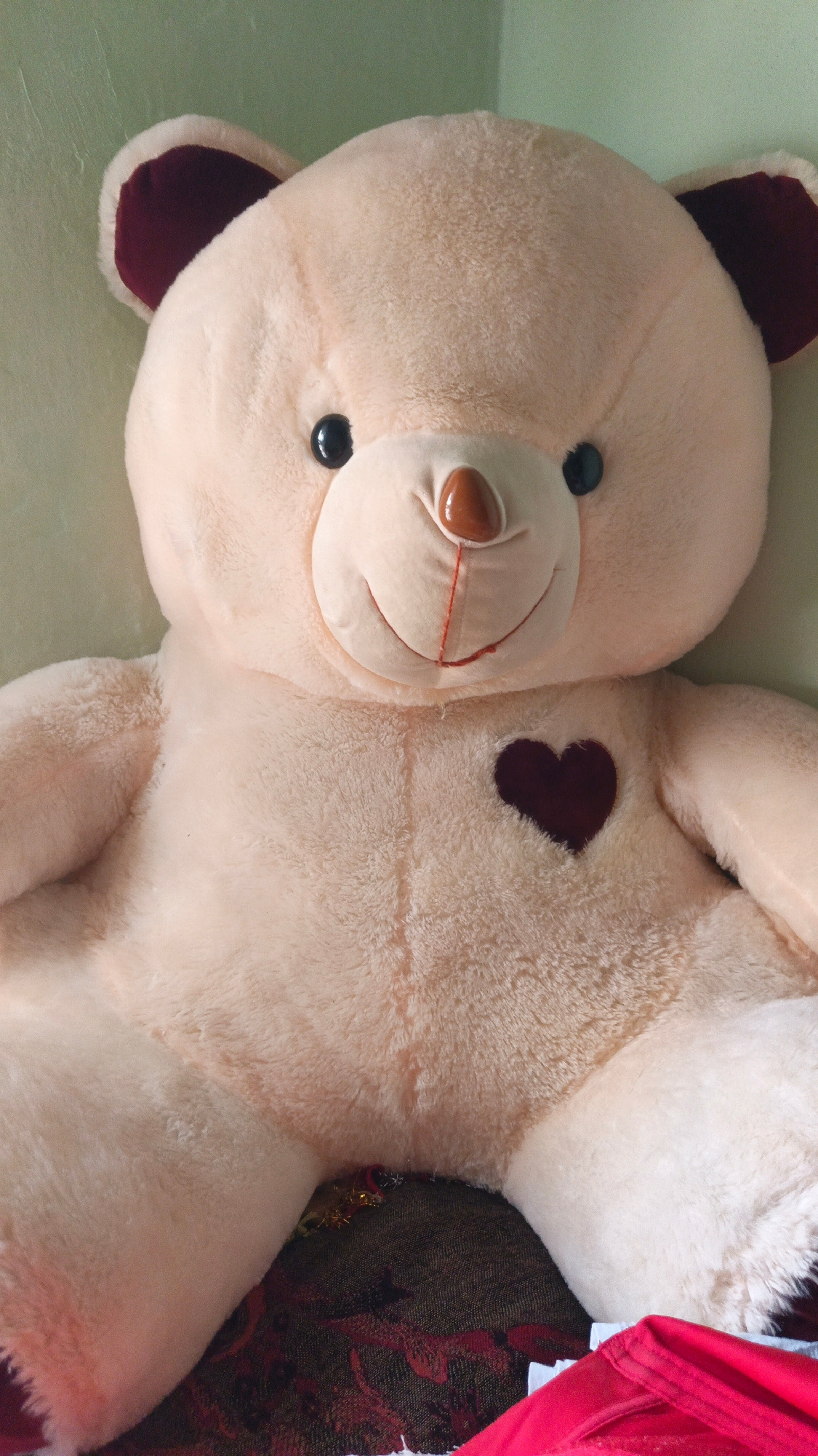 A close-up, front-facing studio shot features a large, plush teddy bear with a pale beige body and dark maroon inner ears. The bear's eyes are round and black, and its nose is a glossy brown. A red, stitched smile adorns its snout, and a dark maroon heart is embroidered on its chest. The bear is positioned upright, appearing to sit on a patterned surface that is partially visible at the bottom of the frame. The background is a plain, light green wall. The lighting is soft, creating no harsh shadows, and the focus is sharp on the teddy bear.