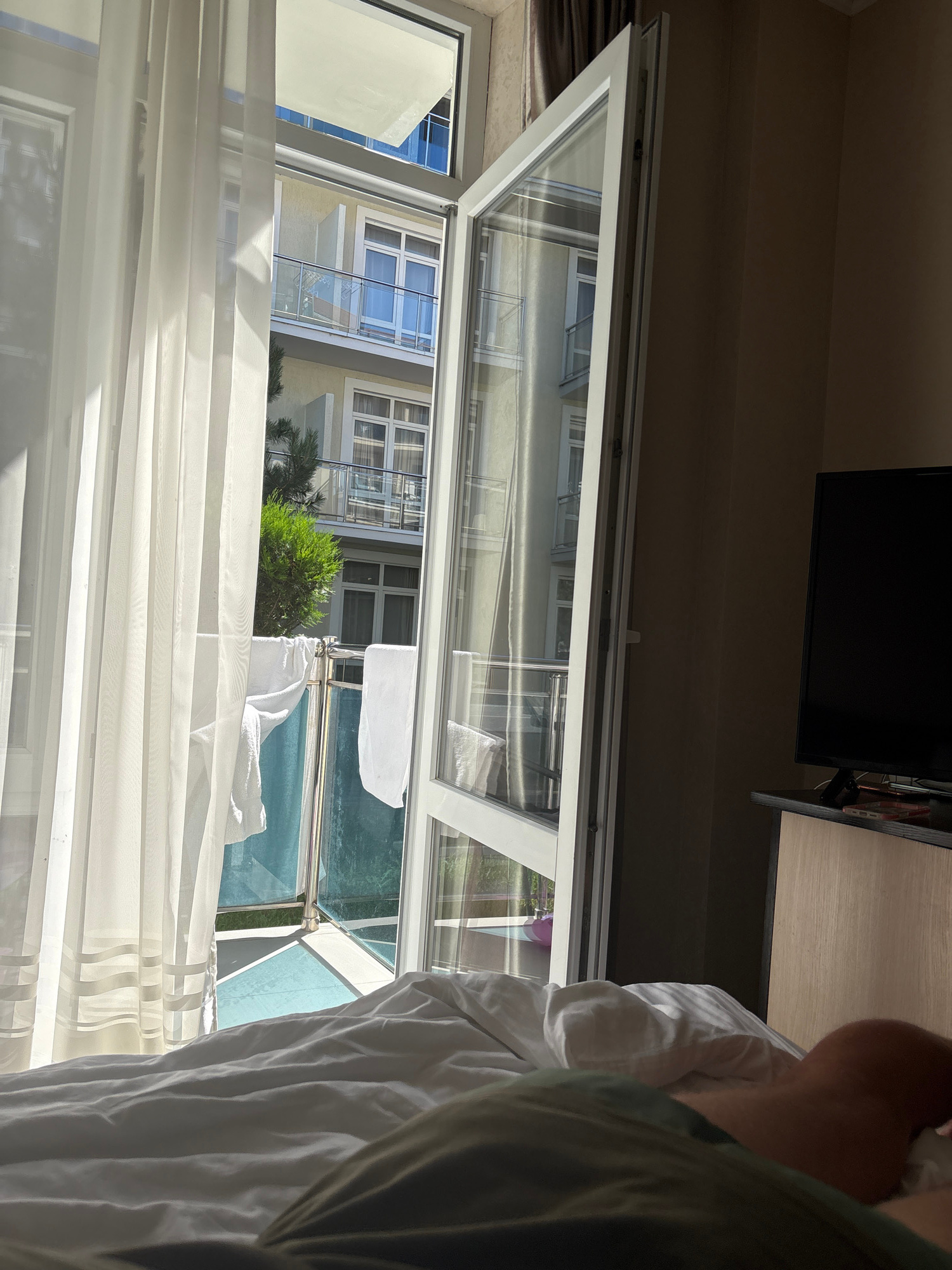 The image shows a view from inside a room, looking out an open window. The room appears to be a bedroom, with white bedding visible in the foreground on the right. The window looks out onto a balcony and then onto an outdoor scene. On the balcony, there are white towels or cloths hanging from a railing, some of which are slightly wet and appear to be drying in the sun. Beyond the balcony, there is a building with multiple floors and many windows, suggesting a hotel or apartment complex. A small green bush is also visible between the balcony and the building.
The lighting suggests it is daytime, likely sunny, as evidenced by the bright light and shadows. The overall impression is a casual, relaxed scene, possibly a vacation. The view of the building and balcony implies a temperate climate. No text is visible in the image. The scene is likely set in Anapa, Russia, as indicated by the location context.