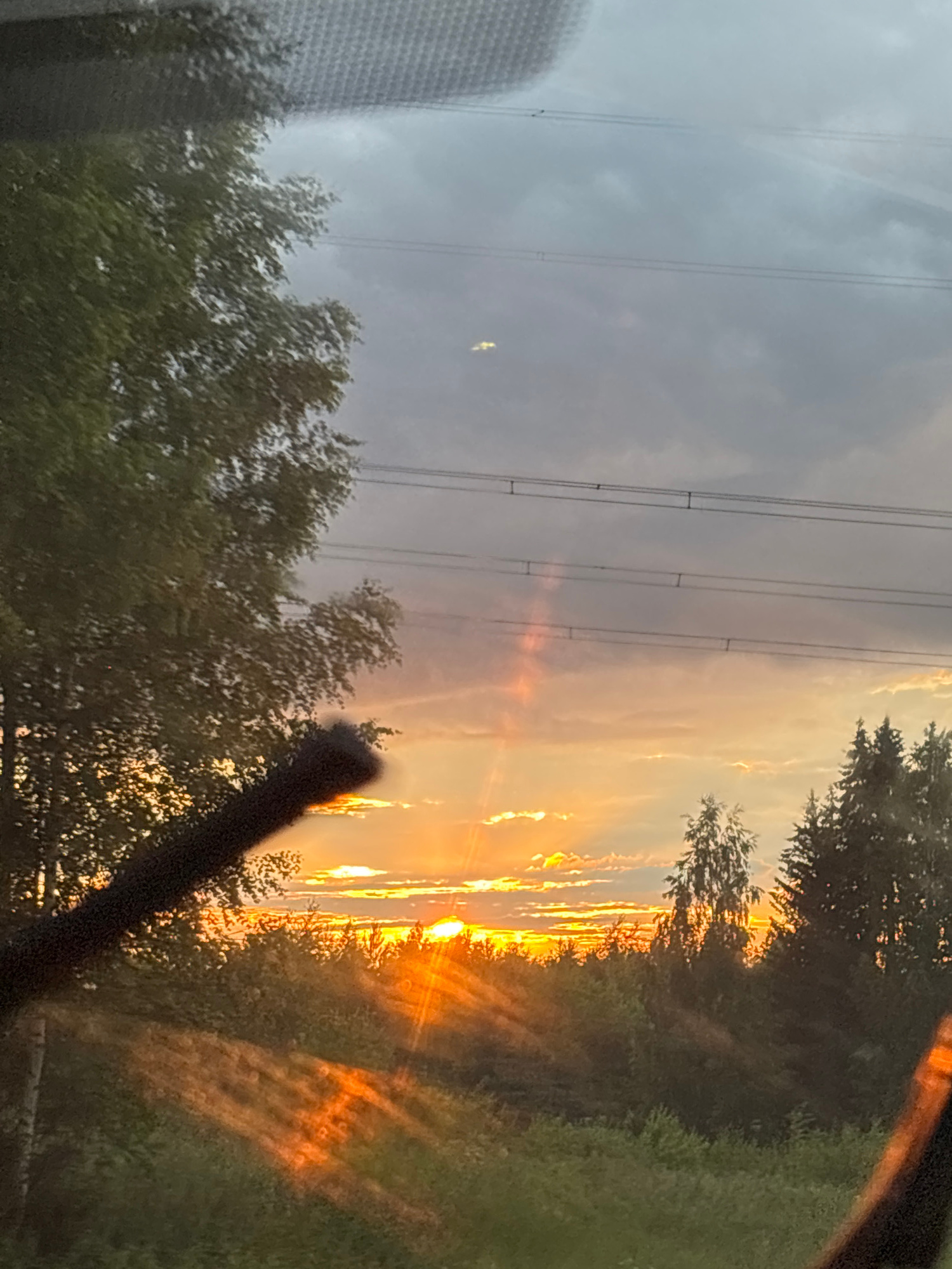 The image depicts a sunset viewed through a car windshield, likely from the perspective of a passenger. The sun is low on the horizon, casting a warm orange glow across the sky and illuminating clouds. Power lines are visible against the sky. In the foreground, a windshield wiper blade is partially obscuring the view on the left, and the edge of the car's dashboard or another part of the interior is visible on the right. The background features a dense line of trees and vegetation, silhouetted against the vibrant sunset. The overall impression is one of natural beauty and a fleeting moment captured during a journey. The specific location of Verkh-Usa, Russia, is not identifiable from the visual elements of the image alone.