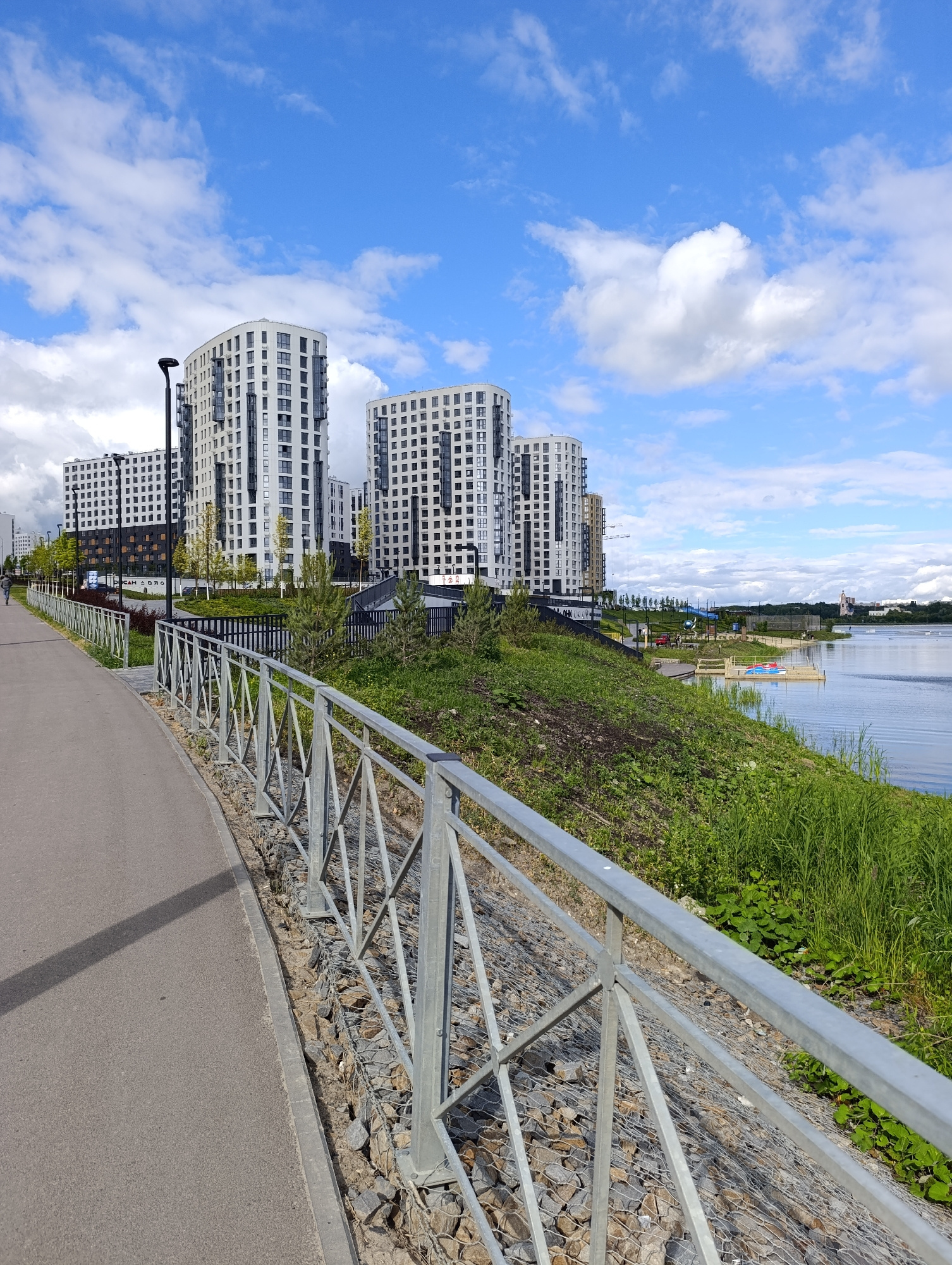 A picturesque scene unfolds along a waterfront promenade, likely in Penza, Russia. The image captures a bright, partly cloudy day, with the sun casting shadows on the asphalt path. In the foreground, a modern metal railing with a geometric design lines the path, separating it from a steep, grassy embankment. The embankment is dotted with scattered rocks and small green bushes, leading down to the calm, reflective surface of a body of water.
Several contemporary, multi-story apartment buildings with white facades and numerous windows dominate the background. Their curved designs suggest modern architecture. A lone figure walks along the path, adding a sense of scale and human presence to the urban landscape. Further along the waterfront, a wooden platform with colorful inflatable structures suggests a recreational area. In the distance, a church with a pink spire peeks above the treeline, providing a historical architectural element amidst the modern development. The overall impression is one of a well-developed urban waterfront, blending modern residential living with leisure and natural beauty.