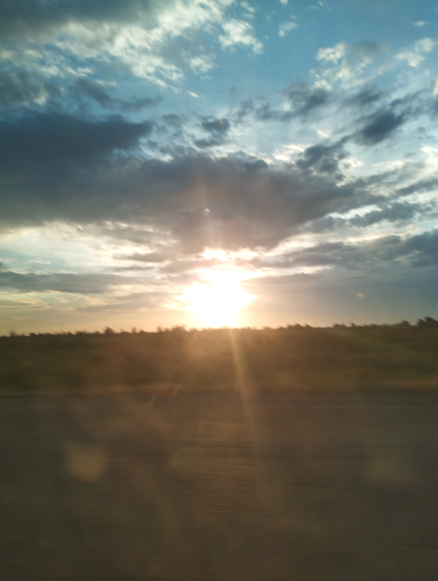 A horizontal, eye-level, medium-shot photograph captures a hazy sunset over a flat, grassy plain with a treeline on the horizon. The sky is filled with a mix of light and dark clouds, with the sun bursting through the center, creating a bright, diffused glow and visible light rays. The foreground is a blur of dark asphalt with horizontal lines, suggesting motion, possibly from a vehicle. The overall lighting is warm and golden, characteristic of sunset. The location is identified as Elabad, Uzbekistan.