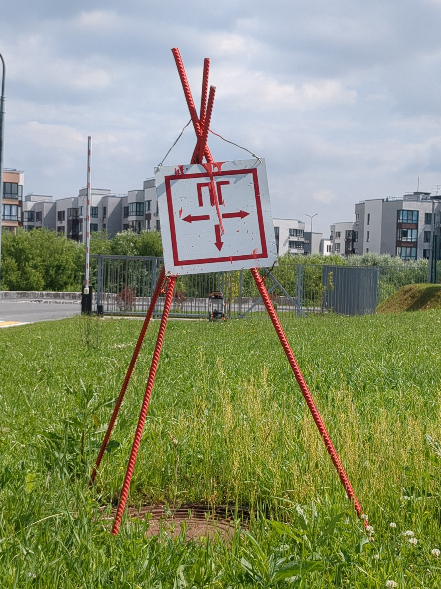 The image depicts a temporary directional sign, possibly indicating construction or a change in route. The sign is a white square with a red border and a red diagram featuring arrows pointing left, right, and down. It is mounted on a tripod-like structure made of red rebar. The sign is situated in a grassy area with tall weeds. In the background, there are modern apartment buildings, a metal fence, and a road with a bollard. The sky is partly cloudy. The location appears to be an urban or suburban area, given the presence of apartment buildings and infrastructure. There are no people visible in the image. The scene appears to be set in an outdoor environment, possibly a park or an undeveloped plot of land within a developed area.