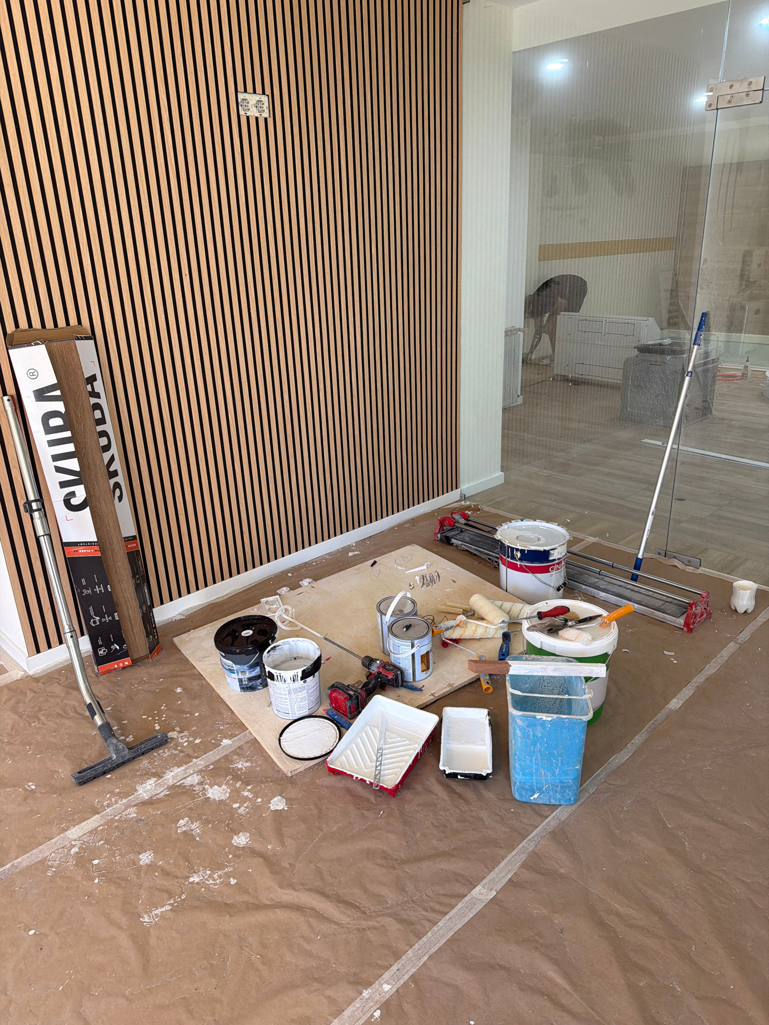 This image depicts a room undergoing renovation or construction, specifically located in Gondomar, Portugal. The scene is characterized by unfinished work and tools commonly associated with painting and tiling.
The walls are partially covered with vertical wooden slats, suggesting a modern design element. On the left side, a large cardboard box with the brand name "SKUDA" is propped against the slatted wall. Next to it, a silver pole with a brush attachment, likely a vacuum cleaner extension, lies on the floor.
The floor is covered with brown protective sheeting, splattered with white paint in various locations, indicating active painting work. Several paint cans are present, including a large white bucket with blue and red accents, another white can with a blue lid, and a smaller black can. Paint rollers, paint trays, and brushes are scattered around, some on a wooden board. A tile cutter is also visible on the floor.
In the background, through a glass partition, another part of the building is visible. This area appears to be a hallway or a larger open space with flooring and some furniture. There are also signs of ongoing work in that area, including what appears to be a person in the distance and a large grey bin. A long-handled mop or squeegee leans against the glass partition.
The overall impression is one of a work in progress, with the necessary tools and materials laid out for painting and possibly tiling. The lighting suggests it is daytime, and the environment is indoors.