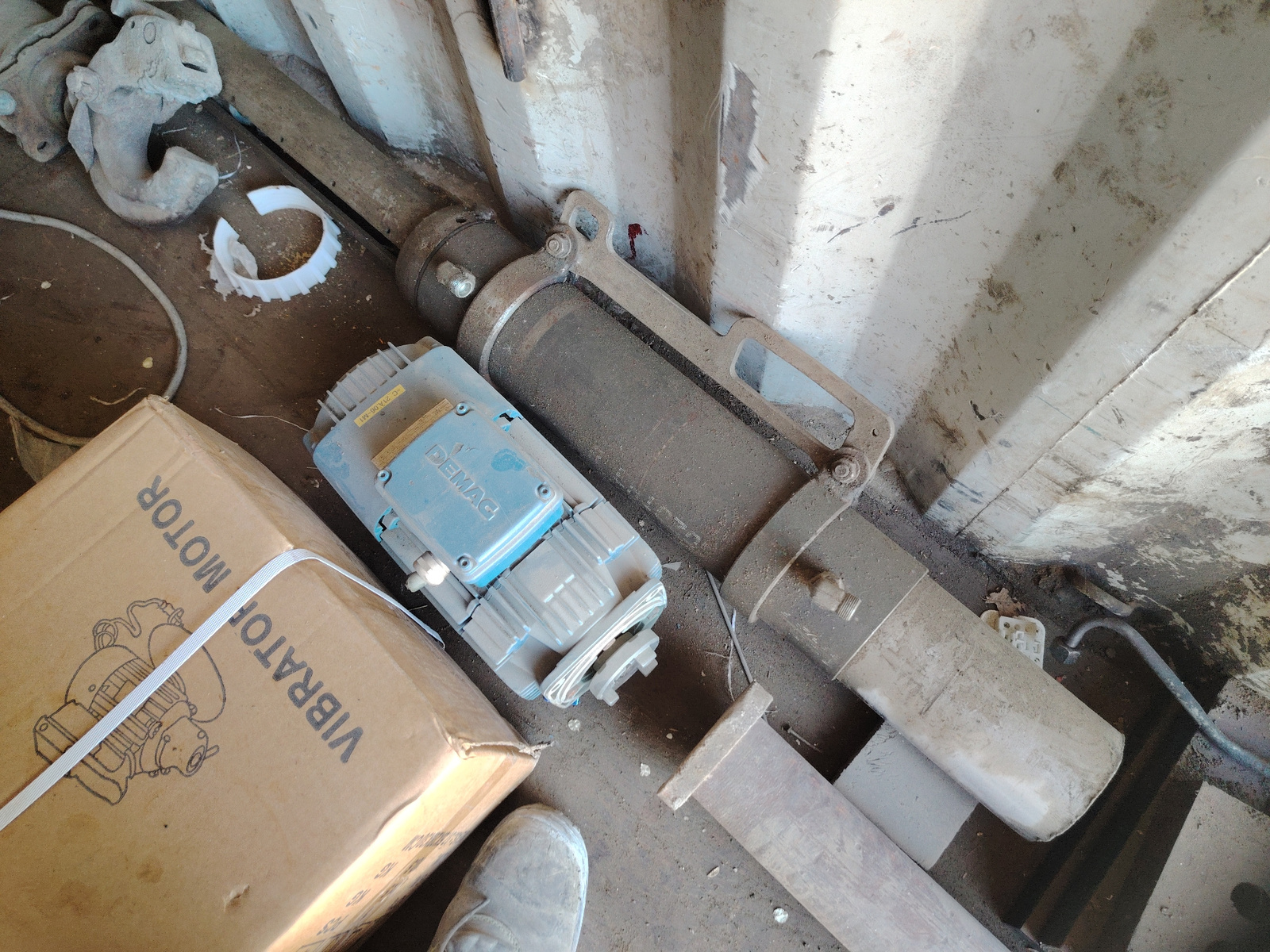 In an indoor setting, possibly a workshop or storage area, several industrial items are visible on a dirty, concrete floor. A large, dusty, cylindrical object with metal fittings rests horizontally. To its left, a blue electric motor with the brand name "DEMAG" is partially obscured by a cardboard box. The box, sealed with white strapping, displays the words "VIBRATOR MOTOR" and an illustration of a motor. Other metallic objects, including a heavy-duty clamp and a length of steel, are also present. A person's grey, work boot is visible in the lower left corner, suggesting human presence or recent activity. The walls of the space appear to be made of rough, possibly corrugated metal. The overall scene suggests a utilitarian environment where heavy equipment is stored or maintained.