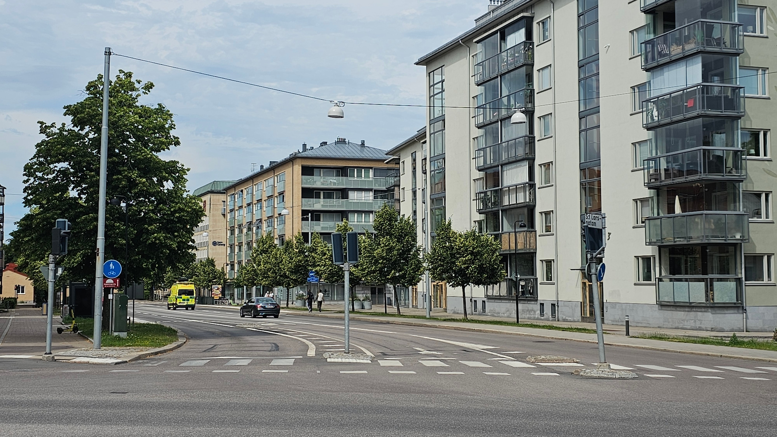 The image shows a street scene in Linköping, Sweden. It is daytime with cloudy weather. The foreground features a road with a pedestrian crossing. To the right, a modern apartment building with multiple balconies is prominently visible. To the left, a traffic light and a street sign are present, along with a yellow ambulance driving on the road. Beyond the ambulance, another apartment building with a yellow facade is visible. Trees line the street, providing greenery. The scene depicts a typical urban environment with everyday activities like driving and walking. There is no apparent emotional tone conveyed. Text visible on the street sign reads "St Lars-gatan".