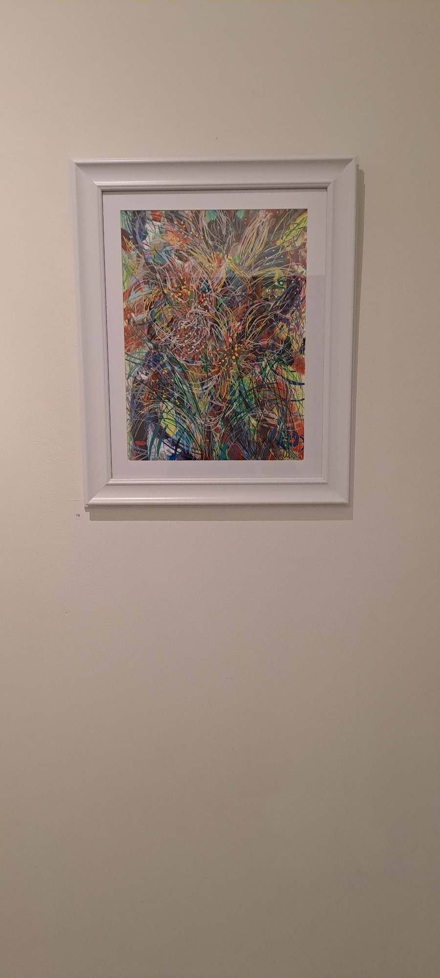 A framed abstract artwork hangs on a plain, light-colored wall. The artwork itself is a vibrant explosion of colors and lines, predominantly featuring shades of green, yellow, red, and blue, with intricate patterns that suggest organic forms or energetic bursts. The frame is a simple, white, rectangular border. Below the artwork, on the wall, a small, discreet number "16" is visible, possibly indicating a catalog or exhibit number. The overall impression is of a piece of art displayed in a gallery or exhibition space. The specific location in Castlebar, Ireland, is not evident from the visual information in the image itself.