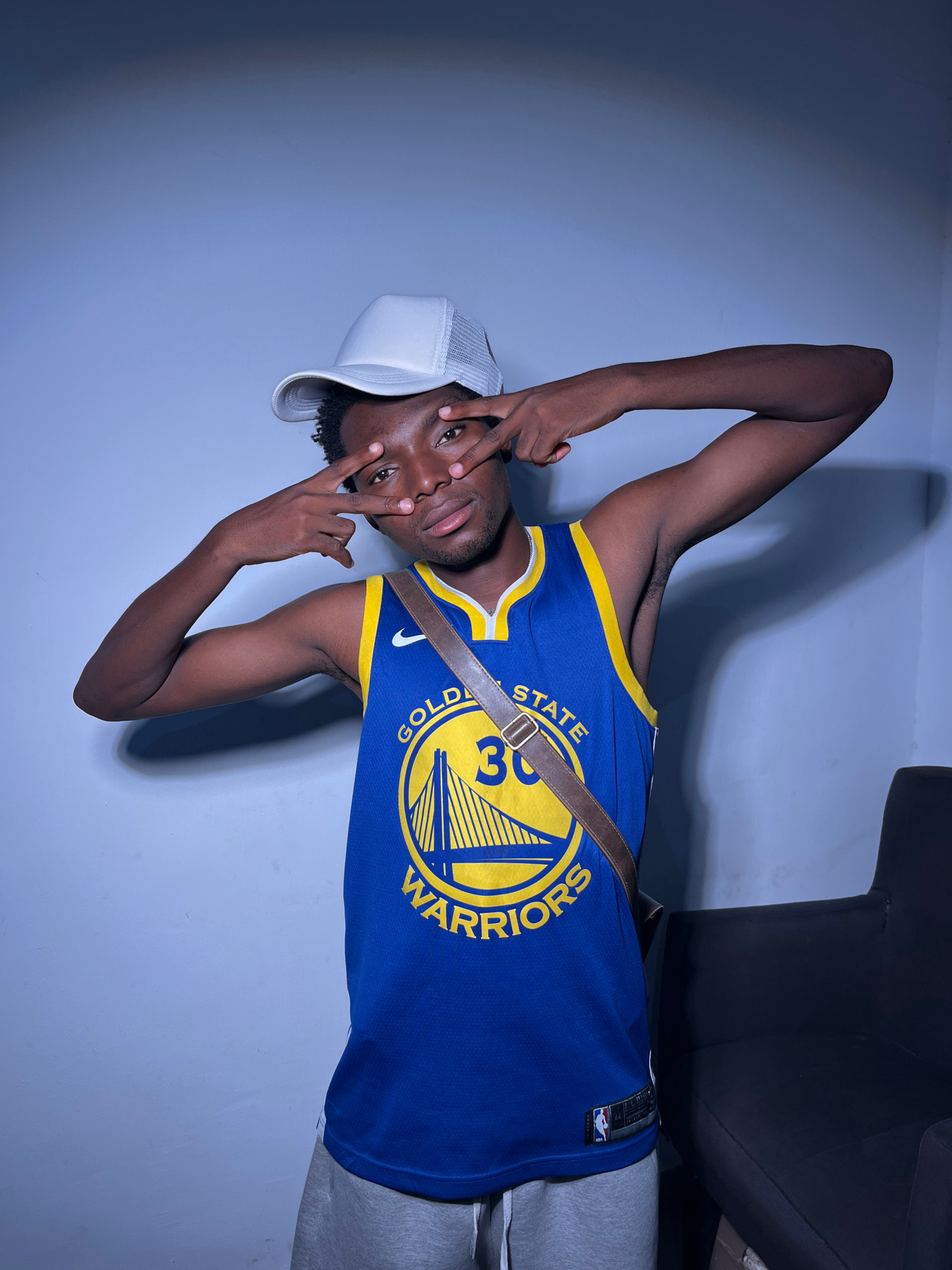 A young Black man is captured in a studio setting, posing with his body turned to the side and his head tilted towards the camera. He is wearing a blue and yellow Golden State Warriors basketball jersey with the number 30 prominently displayed. A brown strap from a bag is slung across his chest, resting on the jersey. He has on a white trucker hat, tilted backward. His expression is direct, with his fingers near his eyes in a gesture that could be playful or intended to obscure his face. The background is a solid, light blue wall, suggesting an indoor environment. The lighting casts a shadow of the man onto the wall behind him. The image is oriented sideways. Visible text includes "GOLDEN STATE" and "WARRIORS" on the jersey, along with a Nike logo and what appears to be a tag on the side of the jersey.