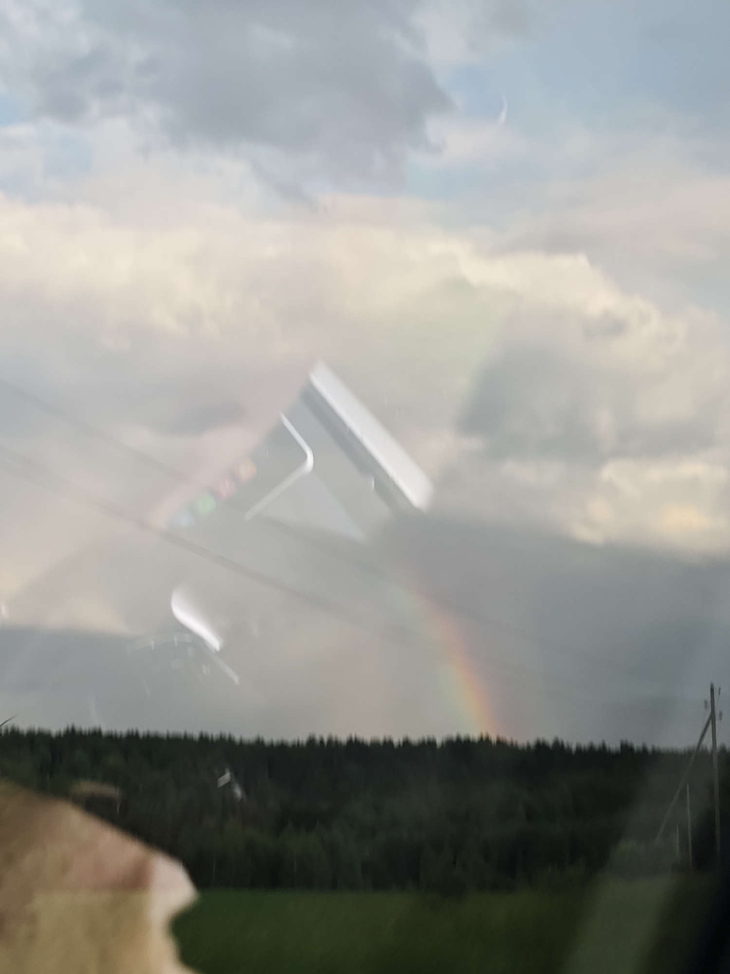 The image displays a reflective surface, possibly a window, superimposed over a natural landscape. The reflection shows a partial view of a device with colored icons, suggesting it's a smartphone or tablet. A faint rainbow arcs across the sky in the background, with dark, dense forest visible on a hill beyond. The sky is filled with dramatic, gray clouds, hinting at a recent or impending rain. A crescent moon is faintly visible in the upper portion of the sky. A wooden utility pole stands to the right of the frame. The lower part of the image shows a blurry green field and what appears to be a portion of a vehicle's interior in the foreground, also reflected on the surface. The location is Foki, Russia.