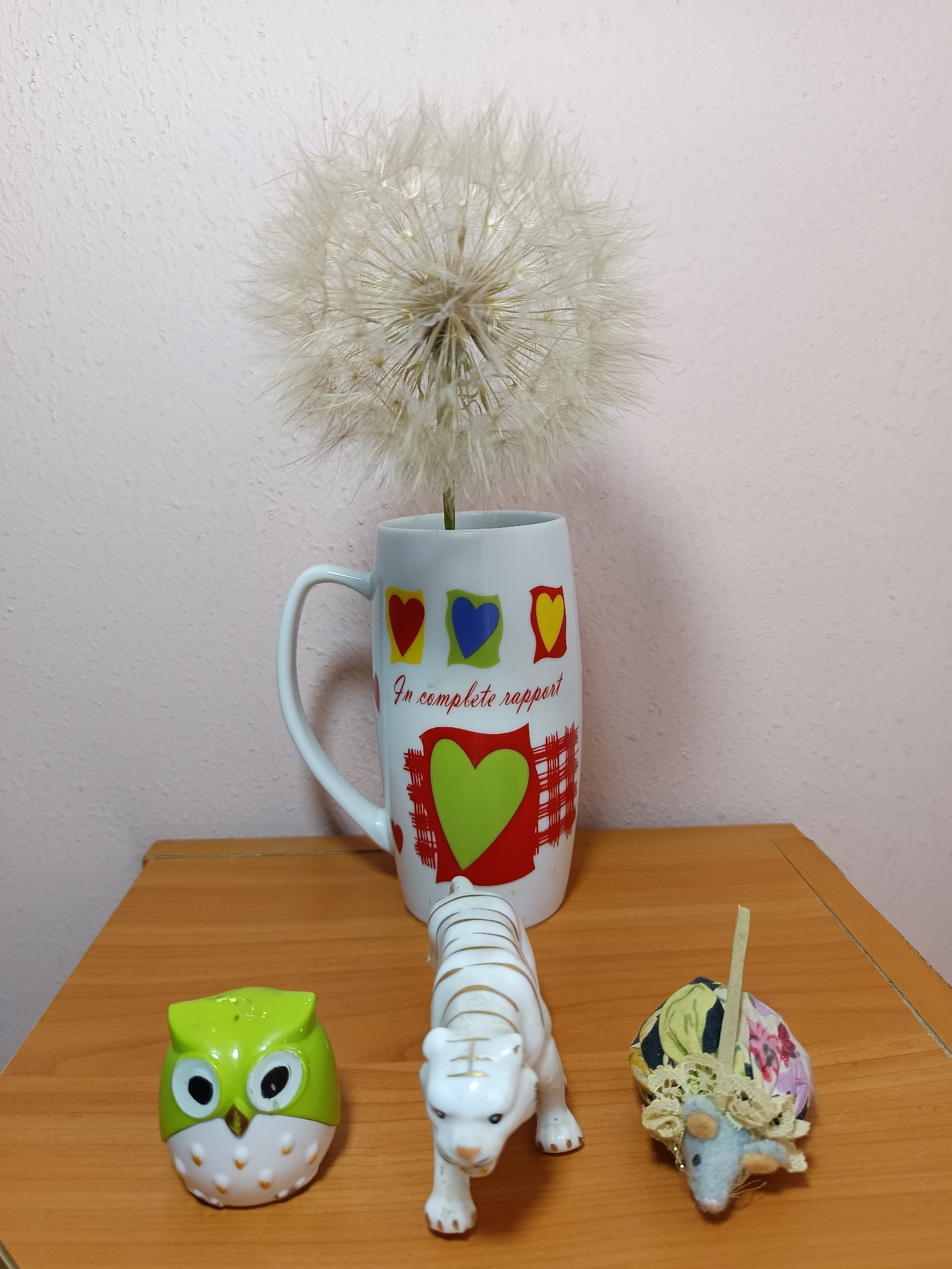 A light pink wall forms the background for a wooden table on which several objects are arranged. On the table, from left to right, are a small, green ceramic owl, a white ceramic tiger with gold stripes, and a small, blue fabric mouse with a colorful fabric dome on its back. Behind these figures, slightly to the right of center, is a white mug decorated with colorful hearts and the text "in complete rapport." A large dandelion seed head is placed inside the mug, with its fluffy white seeds extending outward and upward. The overall lighting suggests an indoor setting, likely a home or office. No people are present in the scene. The location is not specified beyond the general reference to Tyshky, Ukraine.