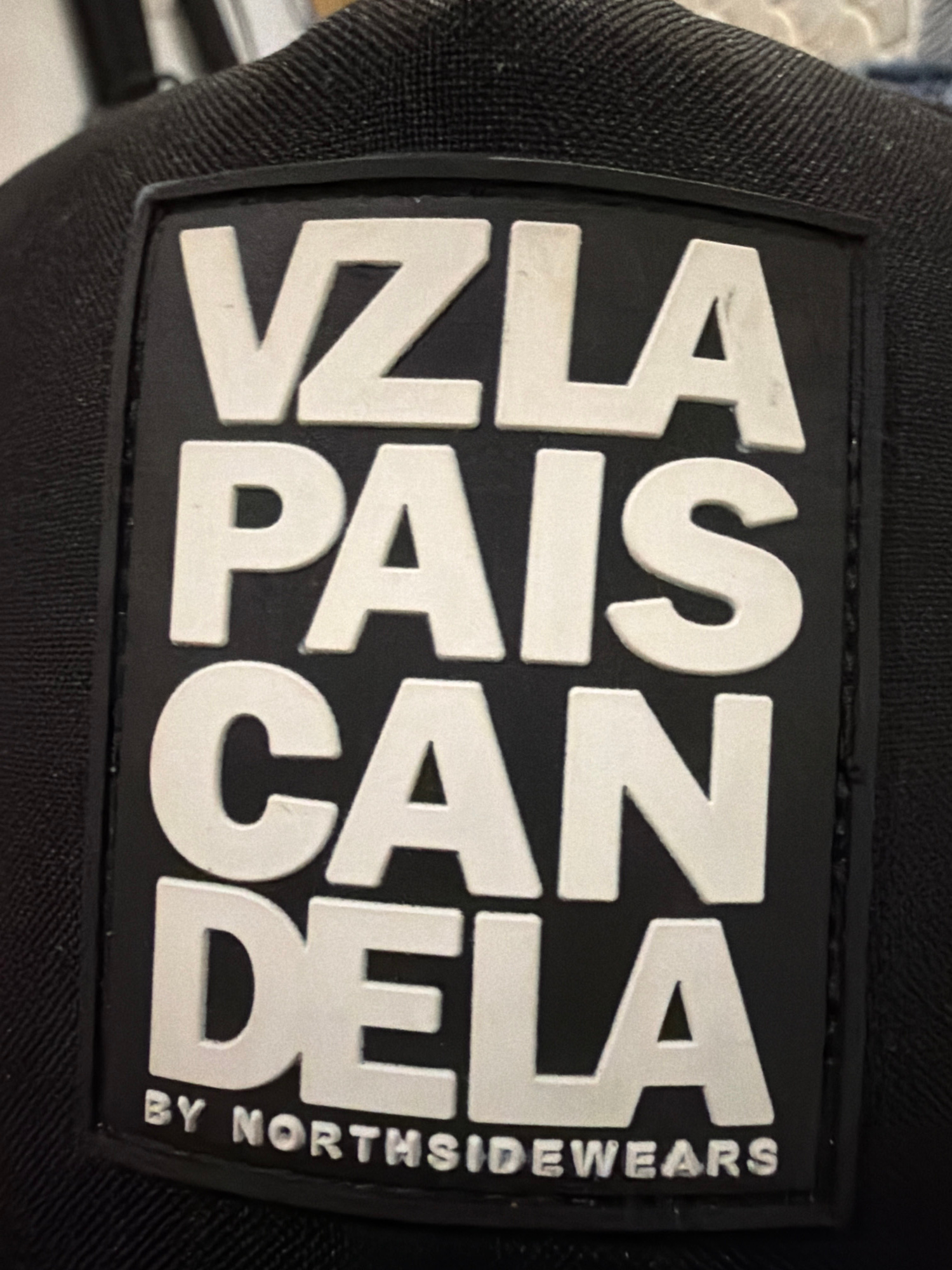 The image shows a black baseball cap with a rectangular patch featuring white text. The patch has the words "VZLA PAIS CAN DELA" arranged vertically, with "BY NORTHSIDEWEARS" written horizontally at the bottom. The cap is black and appears to be made of fabric. The background is dark and out of focus, suggesting the image was taken indoors, possibly in a retail or personal setting. There are no people or animals visible in the image. The image does not provide any information that would allow for its location to be identified in the city of Castelló de la Plana, Spain.