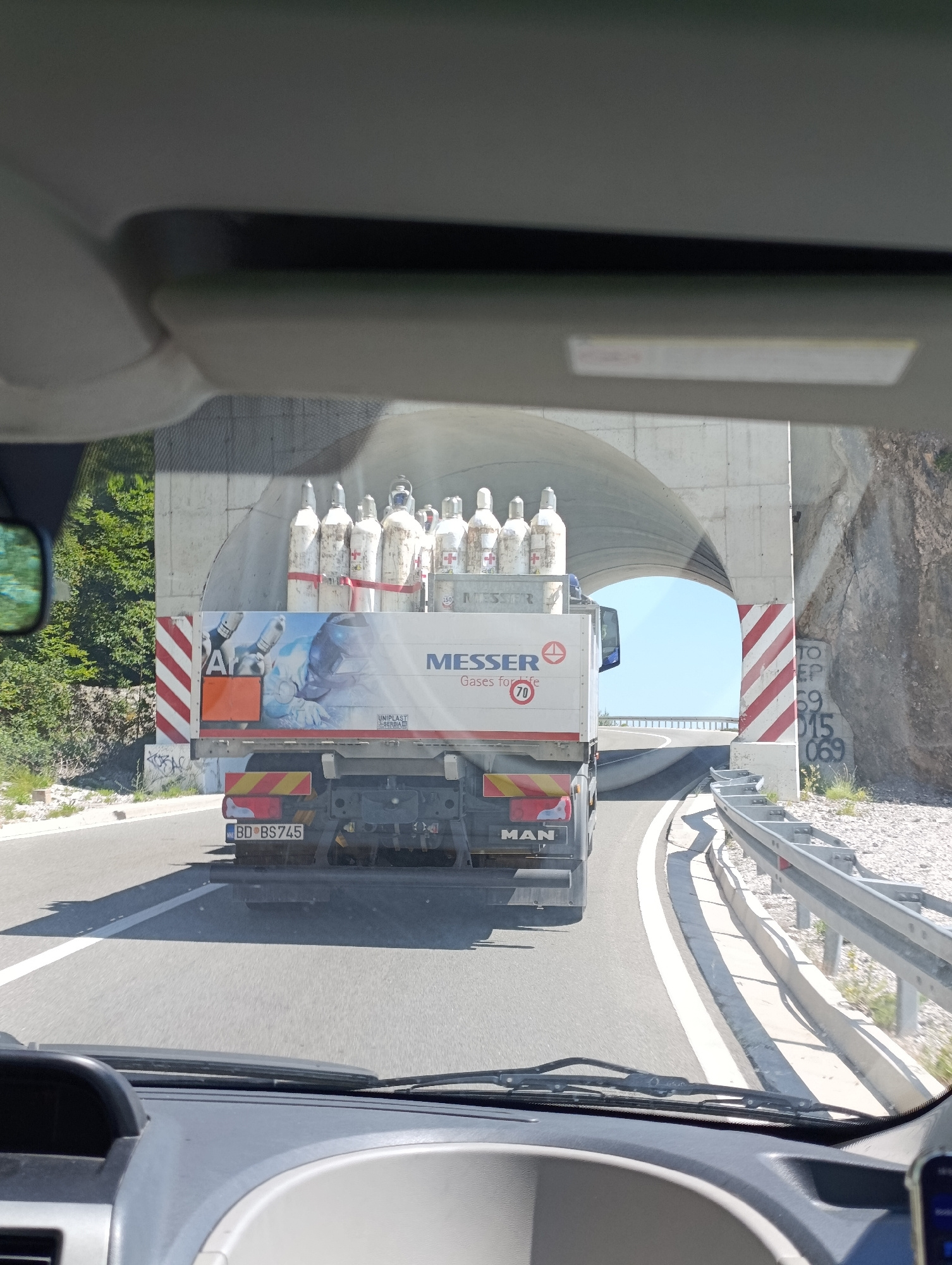 The image captures a view from inside a vehicle, looking out through the windshield at a truck carrying gas cylinders. The truck is driving on a road that leads into a tunnel. The tunnel entrance is made of concrete, and on the right side of the tunnel entrance, there is graffiti on the rock face. The truck has "MESSER Gases for life" written on its side, along with a "70" speed limit sign. The truck's license plate reads "BD BS 745". The surrounding environment appears to be mountainous with trees visible on the left side of the road. The location is identified as Brajici, Montenegro.