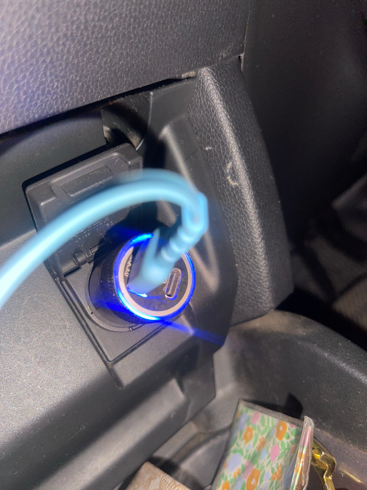 A close-up, slightly angled shot captures a blue USB charging cable plugged into a car's power adapter, which is illuminated with a blue light. The adapter itself is a cylindrical device with a metallic texture and a USB-C port visible. It is inserted into a black car's power outlet, which is located within a textured black plastic console. To the left of the power outlet, a black rectangular cover with some textured detail is visible. In the lower right corner of the frame, part of a compartment within the car is visible, containing a small, colorful object with a floral pattern and a gold-colored metallic edge. The image appears to be taken indoors, likely within a vehicle. The scene does not provide any specific geographic information to indicate the location in Banda Aceh, Indonesia.