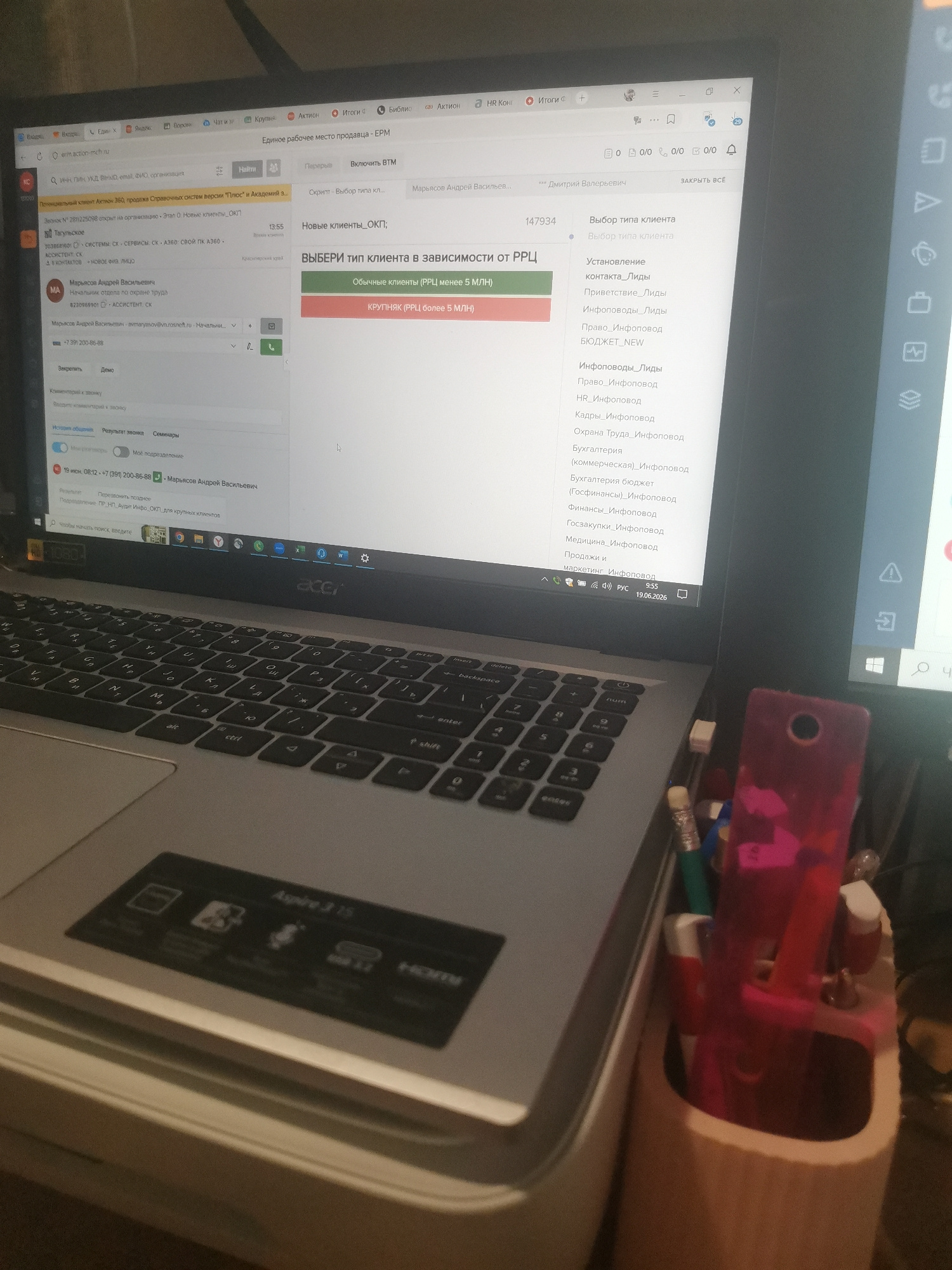 An overhead medium shot shows a laptop computer open and displaying a work-related software interface. The software is in Russian and appears to be a customer relationship management (CRM) system. On the screen, there is a pop-up window with the title "Выбор типа клиента" (Select client type). Below this title, there are two options: "Обычные клиенты (РРЦ менее 5 млн)" (Regular clients (RRC less than 5 million)) and "КРУПНЯК (РРЦ более 5 млн)" (Large client (RRC more than 5 million)). A list of client categories is also visible on the right side of the screen, including "Установление контакта_Лиды" (Establish contact_Leads), "Приветствие_Лиды" (Greeting_Leads), "Инфоповоды_Лиды" (Info-leads_Leads), and many others. The laptop is an Acer Aspire 315. The background is dark and out of focus, but it appears to be an indoor environment, possibly an office or a home desk. A pink object, possibly a pencil holder, is visible to the right of the laptop, containing pens and pencils. The date and time displayed on the laptop screen are 19.05.2026 9:55. There is no indication of the city of Voronezh, Russia in the image.