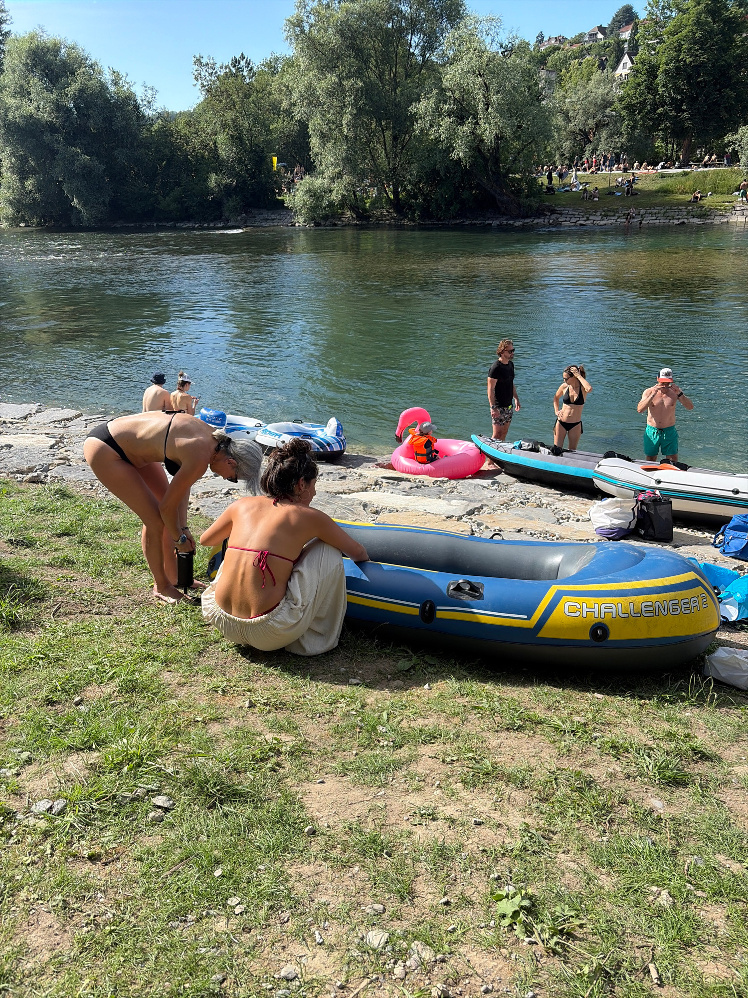 The image depicts a sunny day at the Limmat river in Zurich, Switzerland. In the foreground, two women are on the grassy bank, near an inflatable raft labeled "CHALLENGER 2." One woman, with long gray hair, is bending over and holding a black water bottle. The other woman, with dark hair pulled up, is sitting and facing away from the camera, wearing a pink bikini top and loose white pants.
Several other people are visible near the water's edge. To the right, three individuals are standing by the river: a man in a black t-shirt and patterned shorts, a woman in a black bikini, and a man wearing a white cap and green shorts, who appears to be adjusting his cap. Several inflatable devices, including a pink flamingo float with a child in an orange life vest, are present on the rocky shore. Two other inflatable boats are partially visible, one gray and blue, and another white and blue.
In the background, the river flows gently, and the opposite bank is lined with lush green trees and houses on a hillside, characteristic of the urban landscape of Zurich. More people are scattered along the riverbank on the far side, enjoying the sunny weather. The overall scene conveys a relaxed summer atmosphere.