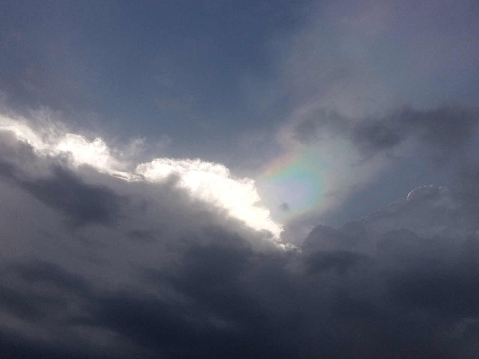 A low-angle, full shot captures a dramatic sky filled with dark, ominous clouds. Sunlight pierces through a gap in the clouds on the left, illuminating a portion of the cloudscape with a bright white glow. Within this illuminated area, a small, iridescent patch of color, resembling a sundog or circumhorizontal arc, is visible. The rest of the sky is dominated by shades of gray and dark blue, suggesting stormy weather. No people or man-made objects are present in the frame.
