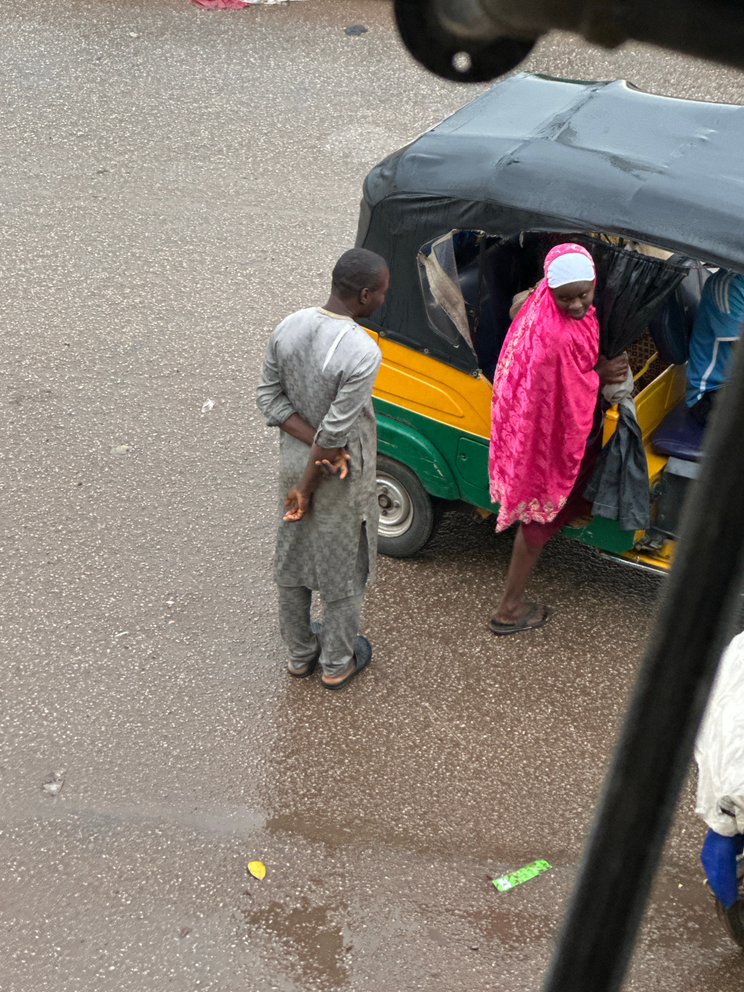 In Bukuru, Nigeria, a man in a grey patterned tunic and trousers stands with his hands behind his back, facing away from a three-wheeled vehicle. A woman, dressed in a bright pink hijab and matching wrap, is partially obscured by the vehicle's roof as she appears to be boarding or disembarking. The vehicle is yellow and green, with a black canopy. The ground is wet and appears to be asphalt, with a yellow leaf visible on the surface. The overall lighting suggests an overcast day.