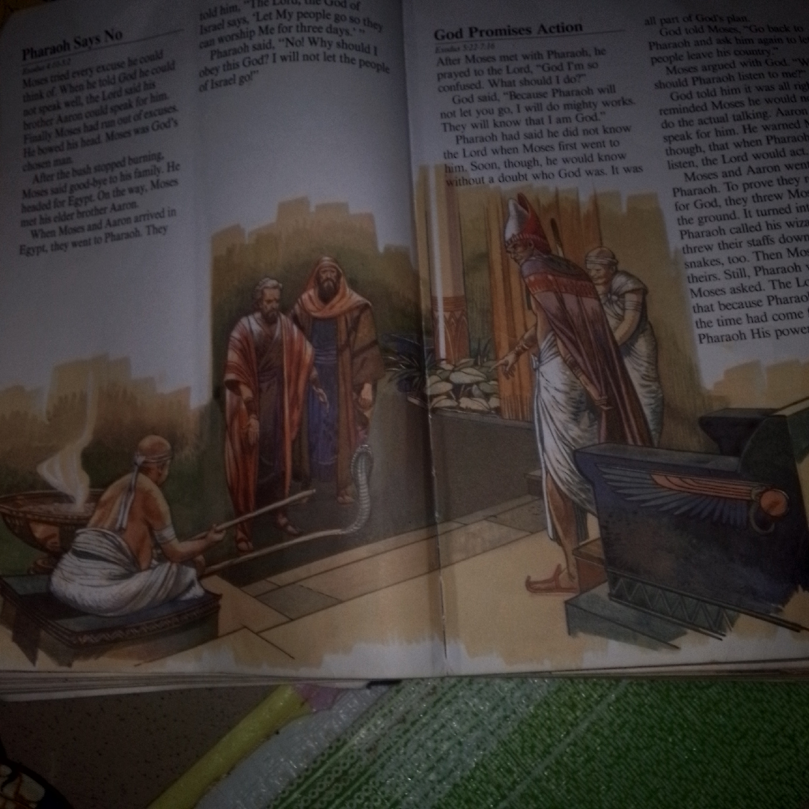 The image depicts an open book with text and illustrations, likely from a religious publication. On the left page, a title reads "Pharaoh Says No," with text below describing Moses and Aaron's interactions with Pharaoh. The illustration shows three figures in ancient attire: two standing and one kneeling by a large, ornate bowl with smoke rising from it. Behind them, a city or structure with brick-like buildings is visible. On the right page, a title reads "God Promises Action," followed by text. The illustration shows two figures in ornate, possibly royal, attire, with one pointing. A coiled snake is also depicted in the foreground of this illustration, and behind the figures are architectural elements that suggest an interior setting. The book lies on a surface that appears to be a table or floor, with other objects, including a yellow marker and a clear plastic bag with green patterns, partially visible at the bottom of the image. There is no indication of Jalingo, Nigeria, in the image or its textual content.