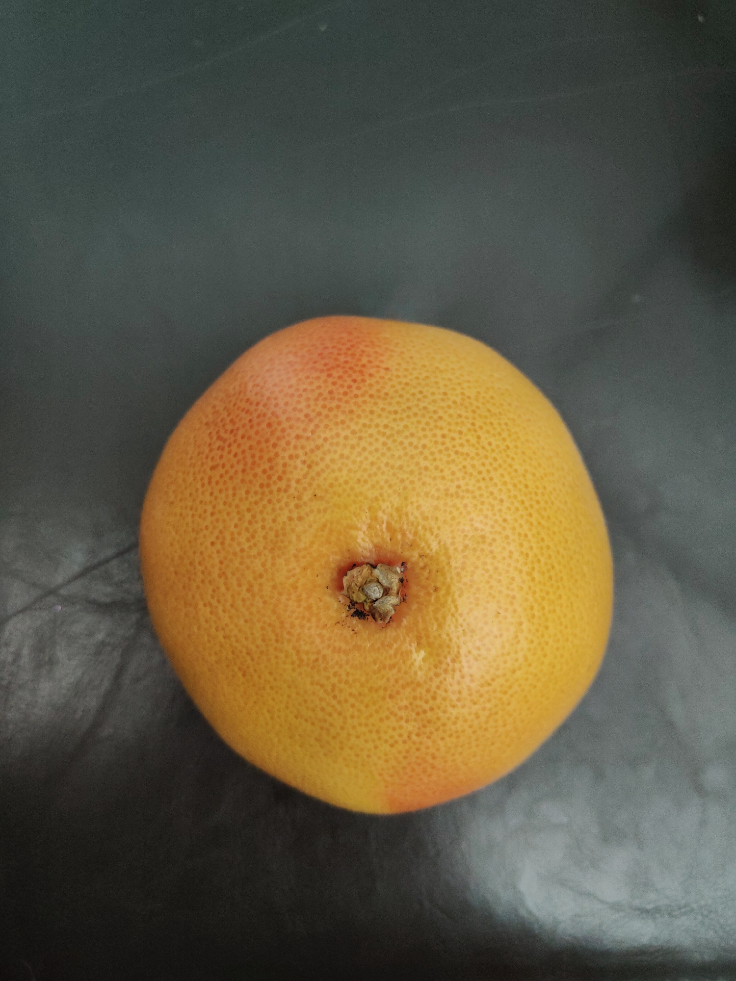 A grapefruit is centered on a dark gray, textured surface. The grapefruit is round and has a textured rind with small pores. Its color is predominantly yellow-orange with a blush of pink on the upper portion. At the top of the grapefruit, there is a small, rough, brown stem scar. The surface it rests on appears to be a matte, possibly synthetic material with subtle lighter gray streaks or reflections, suggesting it might be a countertop or a similar flat surface in a neutral-colored environment. There are no people or discernible objects other than the grapefruit and the surface. The lighting is soft and appears to be coming from above, casting no distinct shadows. The scene is static and devoid of activity. The image is a close-up shot, focusing entirely on the grapefruit and its immediate surroundings.
There is no information in the image that could be used to determine its location in Wrocław, Poland.