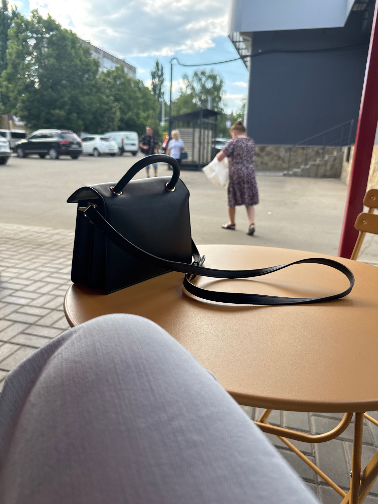 A black handbag rests on a round, beige table. The bag has a structured design with a top handle and a detachable strap. In the background, a person wearing a dark patterned dress walks away from the camera, carrying a white bag. Further in the background, several cars are parked along a street, and a building is visible. The scene appears to be outdoors, possibly a street cafe, with a cloudy sky overhead. The overall impression is casual and urban.