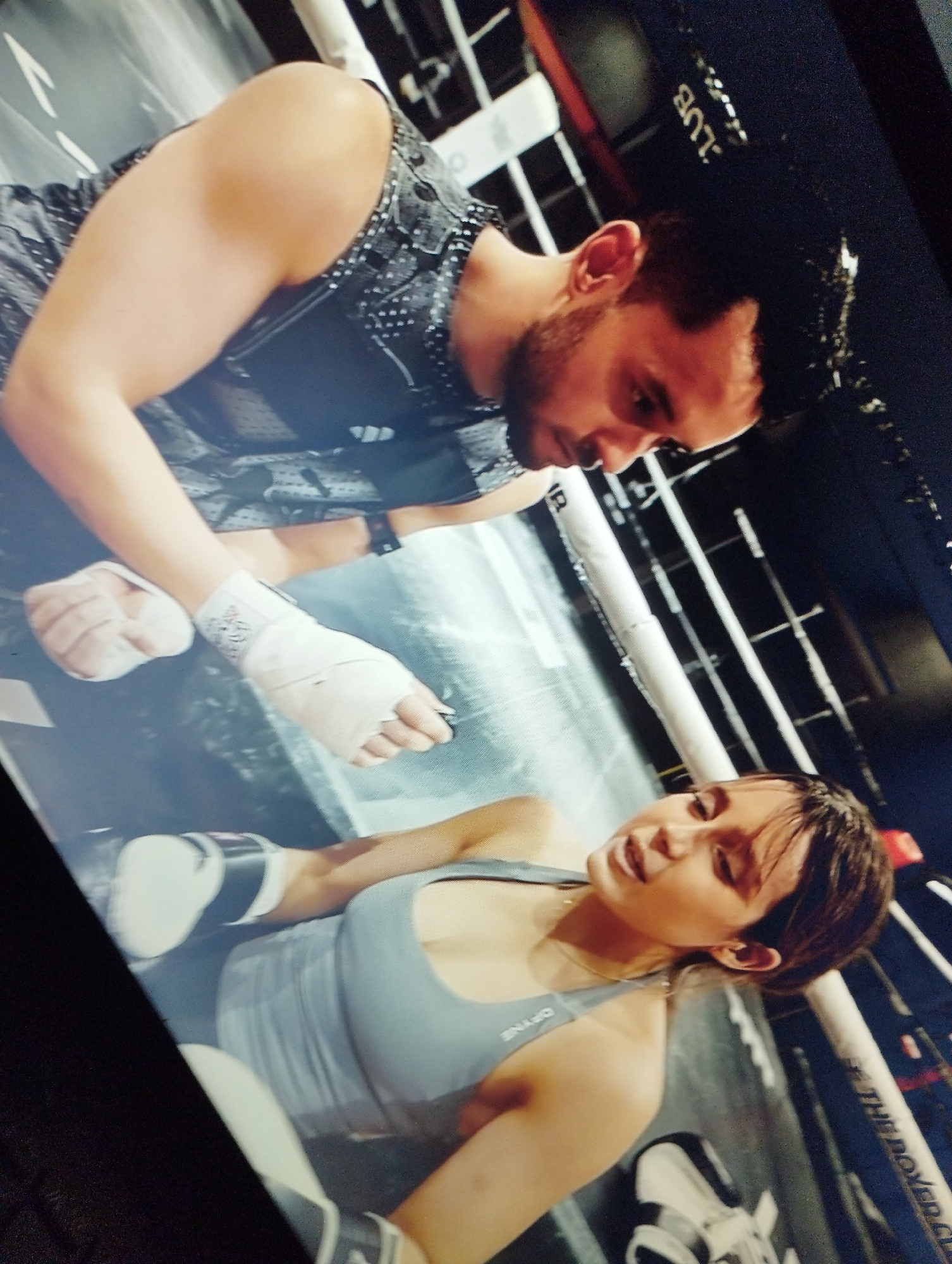 The image depicts two individuals inside a boxing ring, possibly during training or preparation. A man, with short dark hair and a beard, is wearing a dark, patterned tank top and appears to be wrapping the hands of a woman. He is looking down at her hands with a focused expression. The woman, with dark hair and wearing a light grey sports bra, is lying on the canvas of the boxing ring, her eyes are closed or looking down and her mouth is slightly open, suggesting exertion or a moment of rest. Her hands are also wrapped in white tape. White boxing gloves are visible on the floor near her. The background shows the ropes and structure of the boxing ring, with a dark, possibly padded, wall behind it. The lighting is dim, creating a somewhat dramatic atmosphere. There is no information in the image that suggests a specific location within the city of Santa Ana, Venezuela.