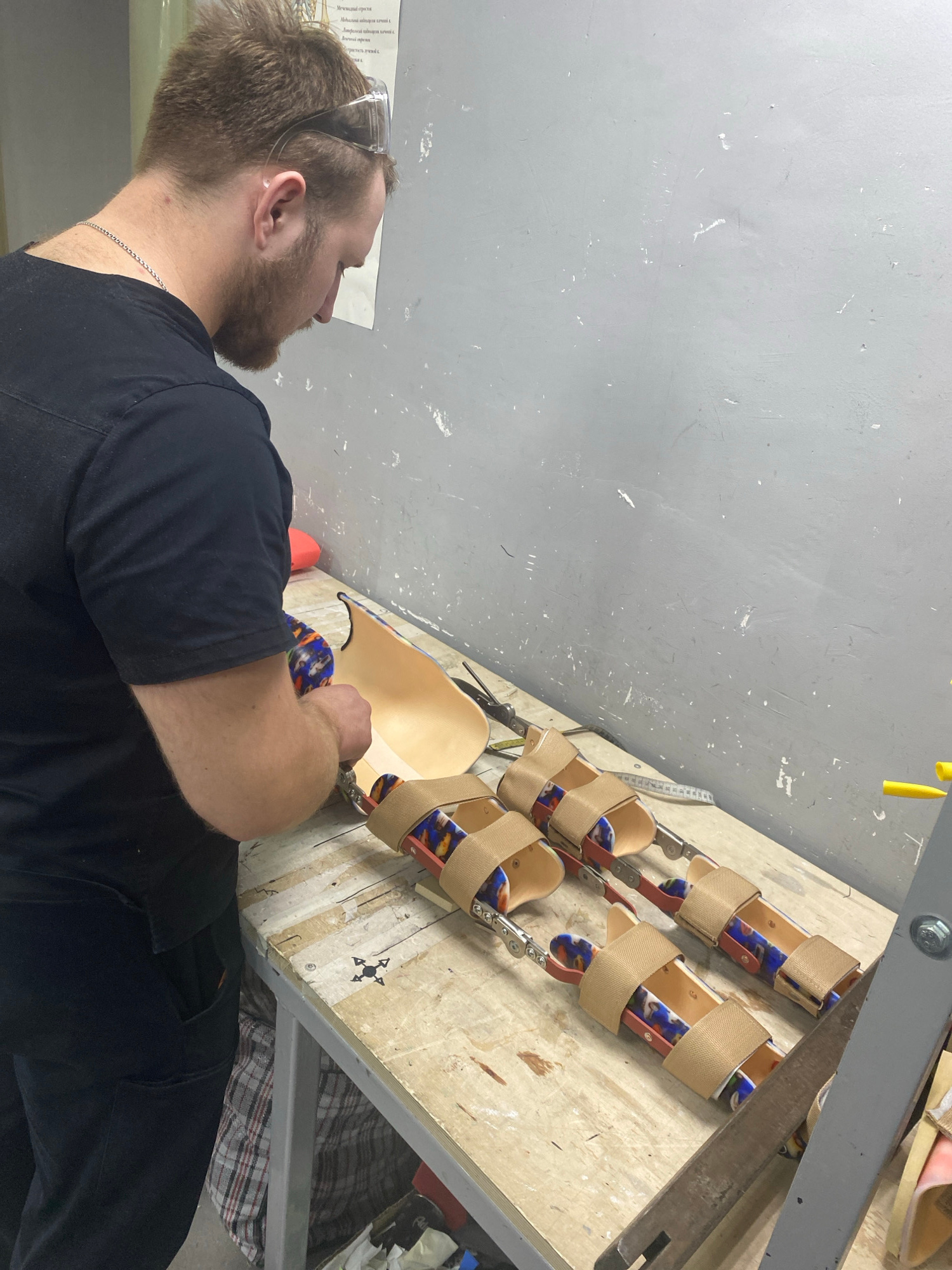 A Caucasian man with a beard and safety glasses is assembling prosthetic legs on a workbench in an unknown location. He is wearing a black t-shirt and black pants. The workbench is made of wood and has various tools and materials on it, including scissors, a measuring tape, and several partially assembled prosthetic leg components with colorful fabric liners and beige straps. The background is a gray, textured wall.