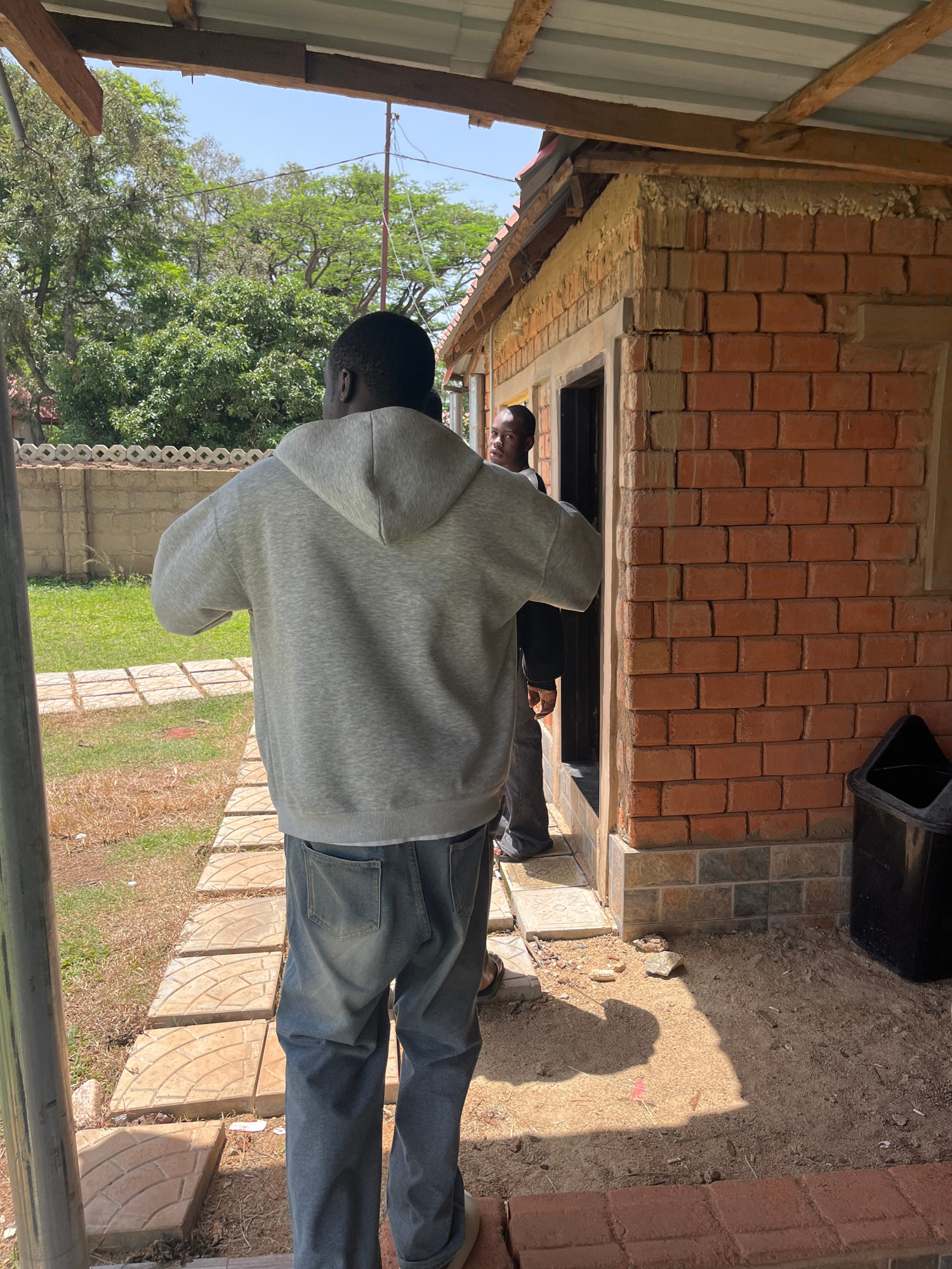 This image shows two people standing outside a building under construction. The person in the foreground is wearing a grey hoodie and blue jeans, with their back to the camera. They are looking into an opening in a brick wall. The second person is partially visible in the opening, wearing a dark shirt. The building is made of red bricks and has a corrugated metal roof. The ground is covered with dirt, sand, and some paving stones. Trees and green vegetation are visible in the background, suggesting a sunny day. The scene appears to be a construction site in Jos, Nigeria, possibly during daytime. There is no visible text in the image.