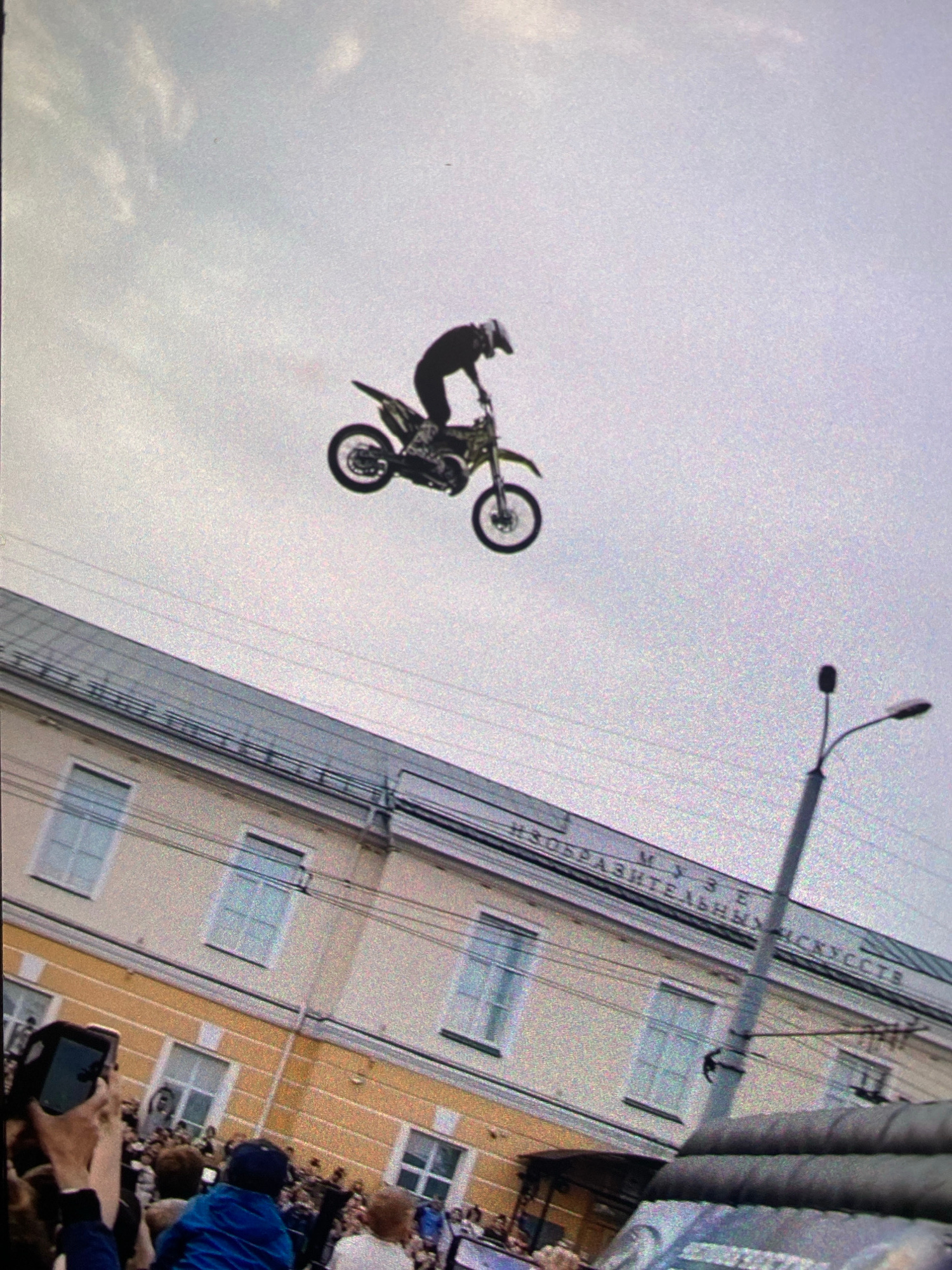 A motocross rider is captured mid-air, performing a stunt with their motorcycle against a hazy sky. Below, a light-colored building with yellow trim stands, its windows reflecting the overcast sky. The building appears to be a museum, with text visible on its facade that reads "МУЗЕЙ ИЗОБРАЗИТЕЛЬНЫХ ИСКУССТВ" (Museum of Fine Arts). A street lamp with a single light fixture is positioned to the right of the building. In the foreground, a crowd of people are gathered, some holding up their phones to capture the event. The lower portion of the image shows a portion of a vehicle, possibly a car or van. The overall scene suggests an outdoor event or performance taking place in a city.