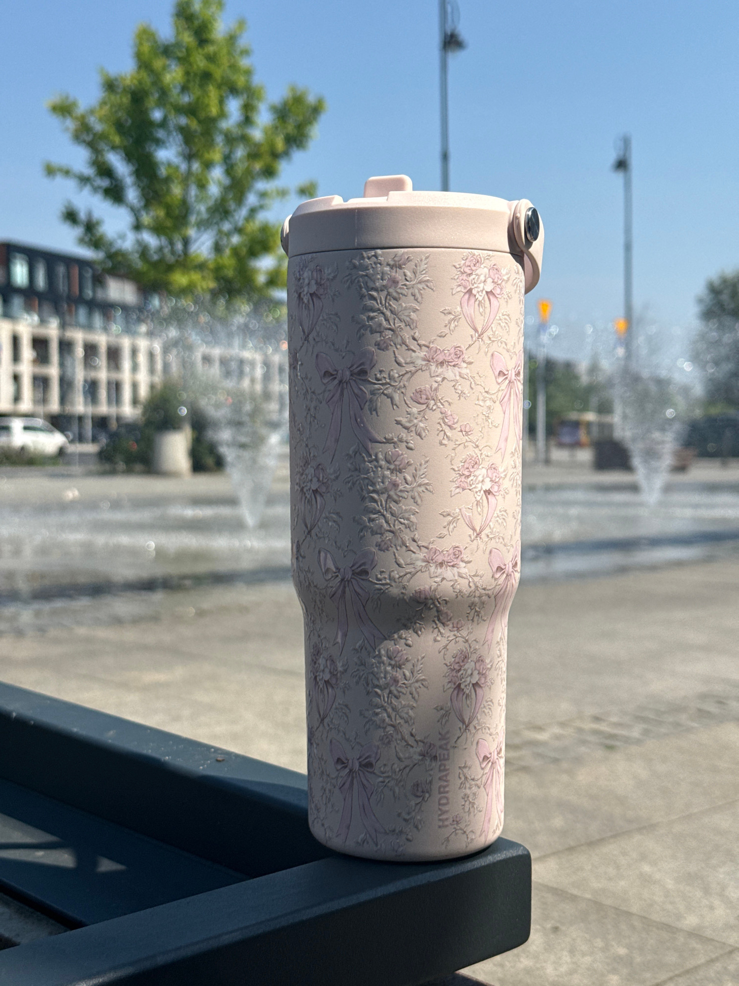 A tall, pink tumbler with a floral and bow embossed pattern sits on a dark blue bench. The tumbler has "HYDRAPEAK" written vertically on its side. In the background, a modern building with many windows and a tree are visible. Further back, a fountain is spraying water. The sky is clear and blue. The scene appears to be in an urban park or plaza. The location is not Warsaw, Poland.