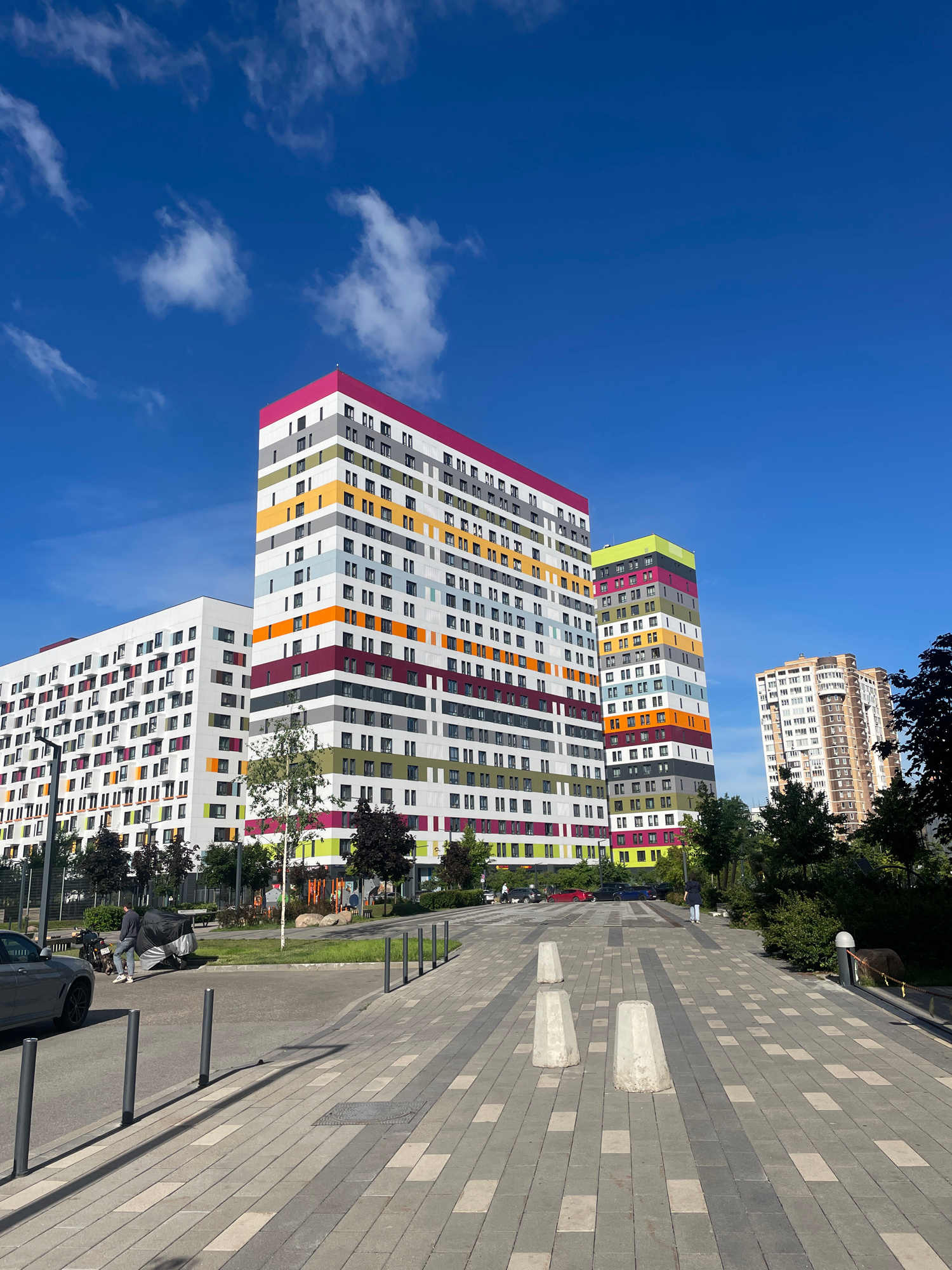 A low-angle, eye-level, full shot captures a modern residential complex against a clear blue sky with scattered white clouds, located in the Shchuchin District of Moscow, Russia. The complex features two prominent, colorful high-rise buildings with a distinctive facade design incorporating bands of white, grey, pink, yellow, orange, green, and burgundy. The building on the left is a long, white structure with colorful window frames. The taller building on the right is adorned with horizontal stripes of various colors. In the background, a lighter-colored, more conventionally designed residential building is visible. The foreground is a paved pedestrian walkway with a grey and beige interlocking paver pattern, lined with dark grey bollards. Three concrete traffic barriers are positioned on the walkway. To the left of the walkway, a grey sedan is parked on an asphalt surface, with a person standing next to a motorcycle covered with a black tarp. A few trees with green foliage are scattered throughout the scene.