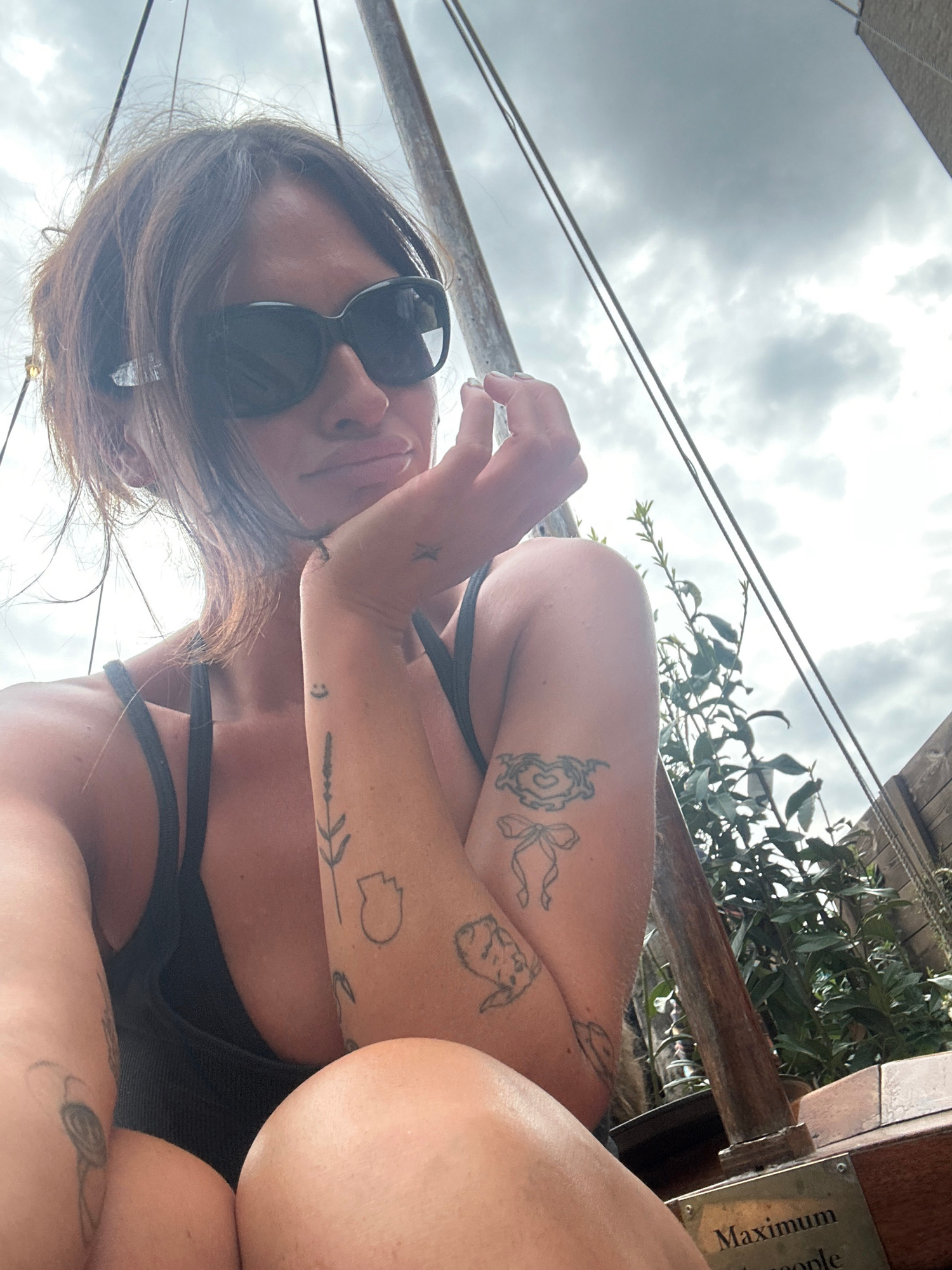 A woman with brown hair and sunglasses rests her chin on her hand, looking towards the camera. She is wearing a black tank top. Her arm is adorned with several tattoos, including a lavender sprig, a smiley face, and a bow. In the background, green foliage and a wooden structure are visible. The sky is overcast. The scene appears to be outdoors, possibly in a garden or park setting. There is a plaque on the wooden structure that reads "Maximum people." The overall lighting suggests it is daytime, but the cloudy sky creates a diffused light. No specific location in London is identifiable from the image.