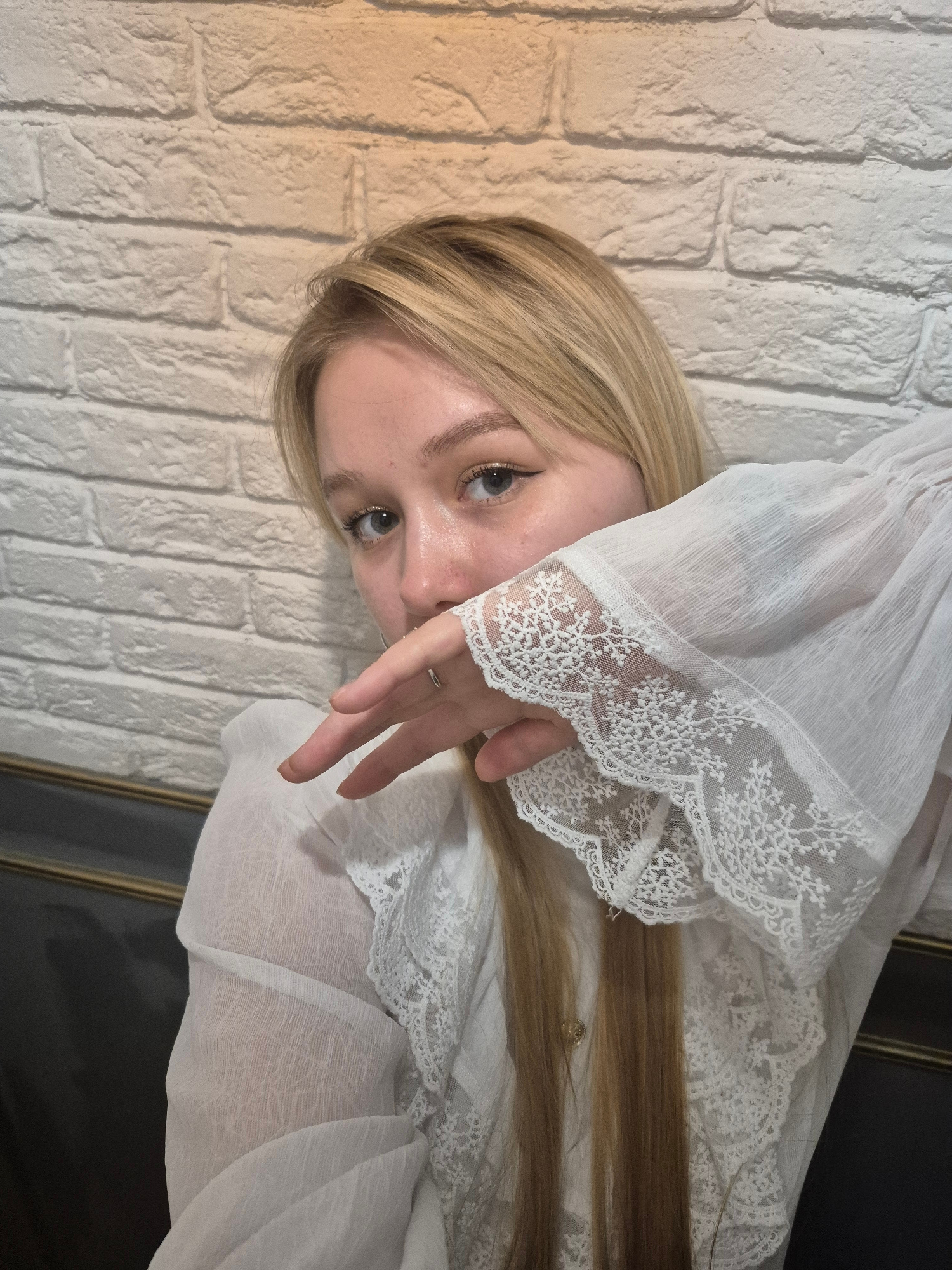 A medium shot captured from a slightly elevated angle shows a young Caucasian woman with blonde hair and light blue eyes. She is wearing a white sheer blouse with delicate lace trim on the sleeves and neckline. Her right hand is raised to her face, obscuring part of her mouth and nose, with her fingers splayed. She is wearing a silver ring on her right ring finger. Her left eye is visible, and she appears to be looking directly at the camera. The background consists of a white brick wall and a dark upholstered surface, possibly a sofa or chair, with gold trim. The lighting suggests an indoor setting, possibly a cafe or restaurant, though no specific location in Chaikovsky, Russia, can be identified from the visual information provided. The overall tone of the image is soft and intimate.