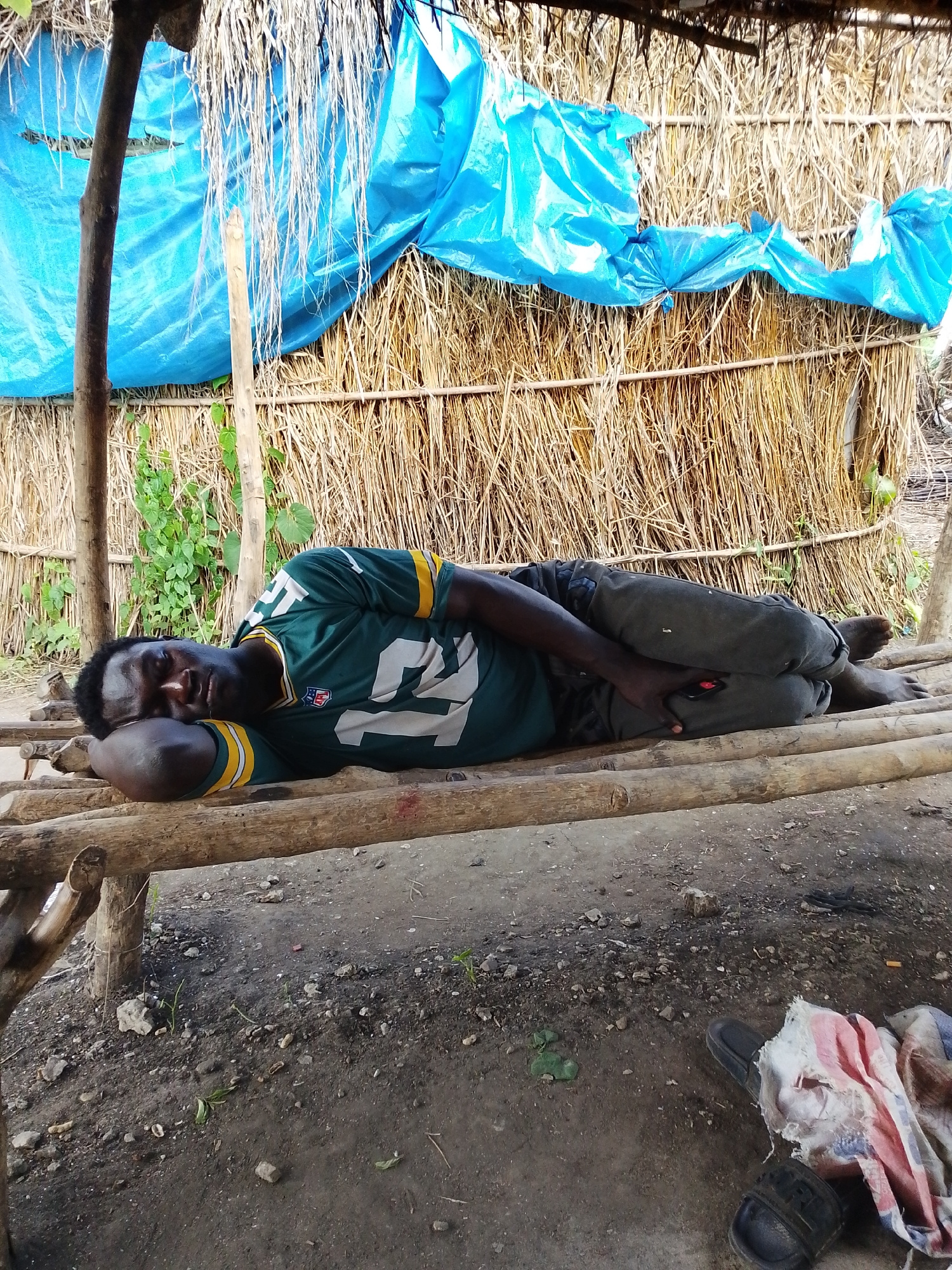 A Black man lies sleeping on a wooden platform, his head resting on his arm. He is wearing a green and yellow football jersey and dark pants. Behind him is a wall made of dried straw, covered in part by a blue tarp. Green foliage is visible to the left of the man, and the ground in front of the platform is dirt with scattered stones and debris. A pair of sandals and a tattered cloth are on the ground to the right. The scene appears to be outdoors in a rural or semi-rural setting, possibly in the city of Otulu, Nigeria.