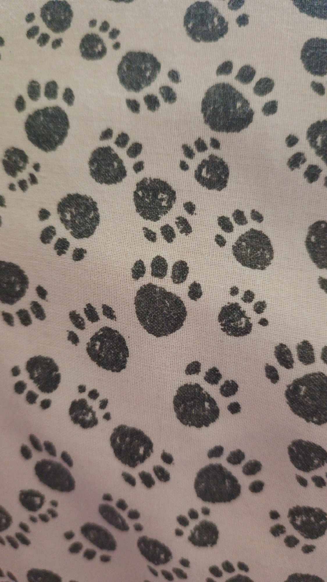 A close-up, vertically oriented shot captures a light pink fabric with a repeating pattern of dark grey paw prints. The paw prints are varied in size, with some appearing larger and more detailed, and others smaller and more abstract. They are distributed across the fabric in an irregular, somewhat scattered fashion. The texture of the fabric is visible, showing a woven pattern beneath the paw prints. The scene appears to be an indoor setting, likely a textile or home furnishing context, with no discernible outdoor or urban elements. The location is Unknown, Unknown.
