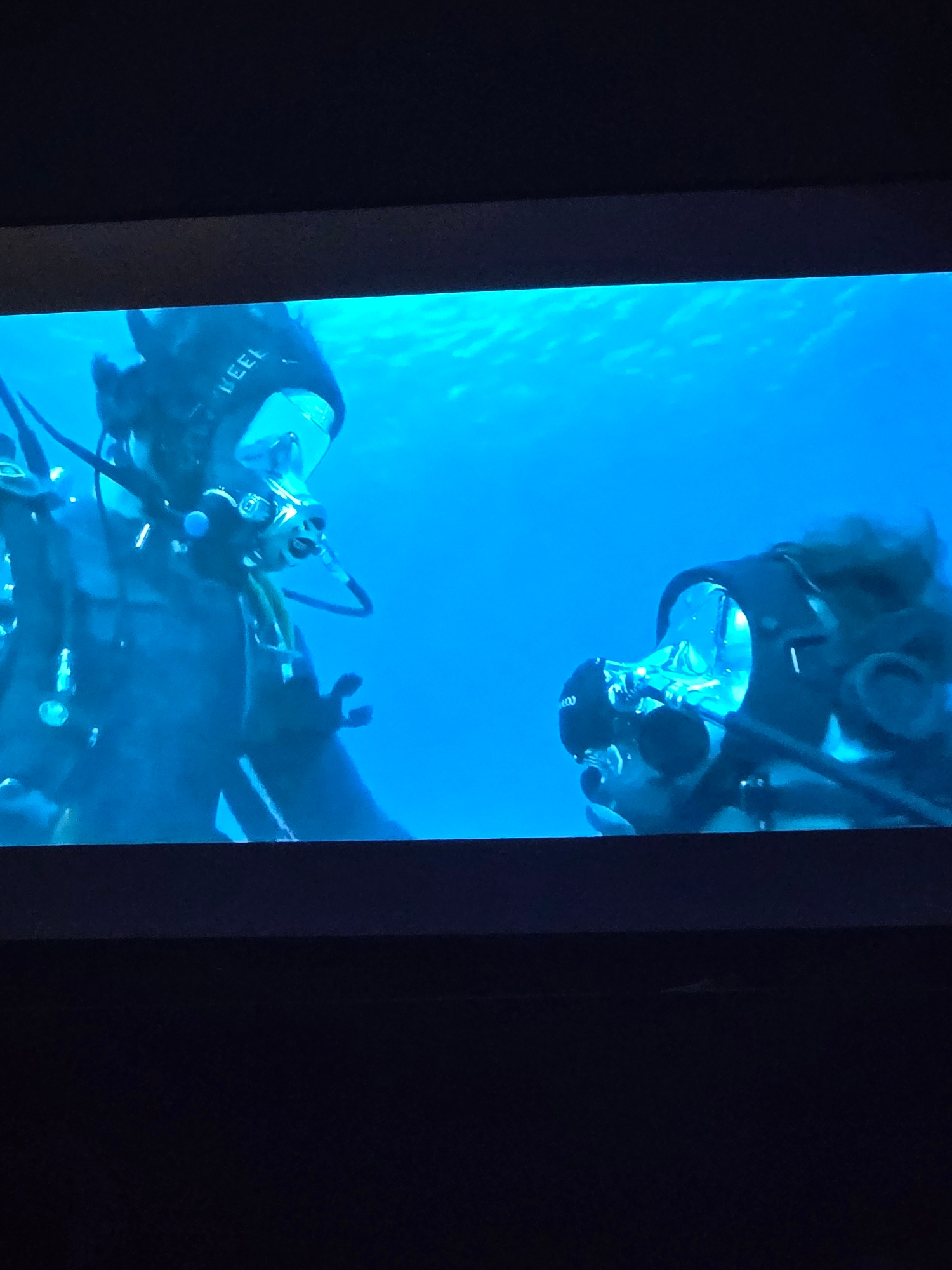 The image shows two divers underwater, illuminated by blue light filtering from the surface. Both divers are wearing black wetsuits and full-face masks, with scuba gear. The diver on the left is closer to the camera, and the word "KEEL" is visible in white lettering on their headgear. They are looking towards the other diver, who is positioned slightly to the right and further away. This second diver's face is obscured by their mask, and their head is tilted as if in conversation or observation. The water is clear and blue, with subtle light reflections visible on the surface above. The scene appears to be taking place in an ocean or a large body of water.
