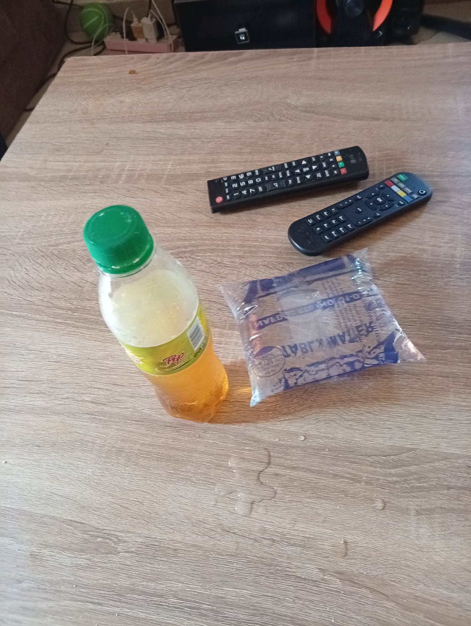 The image depicts a table with several objects on it. Two television remote controls are visible, one above and slightly to the left of the other. In front of the remotes, to the left, is a plastic bottle with a green cap, containing a yellowish liquid. To the right of the bottle, and slightly in front of it, is a small, clear plastic bag filled with a clear liquid. The table surface has a wood grain pattern. The background is out of focus but appears to show some electronic equipment and household items. There are a few water droplets on the table surface. The location in Gura Top, Nigeria is not discernible from the image.