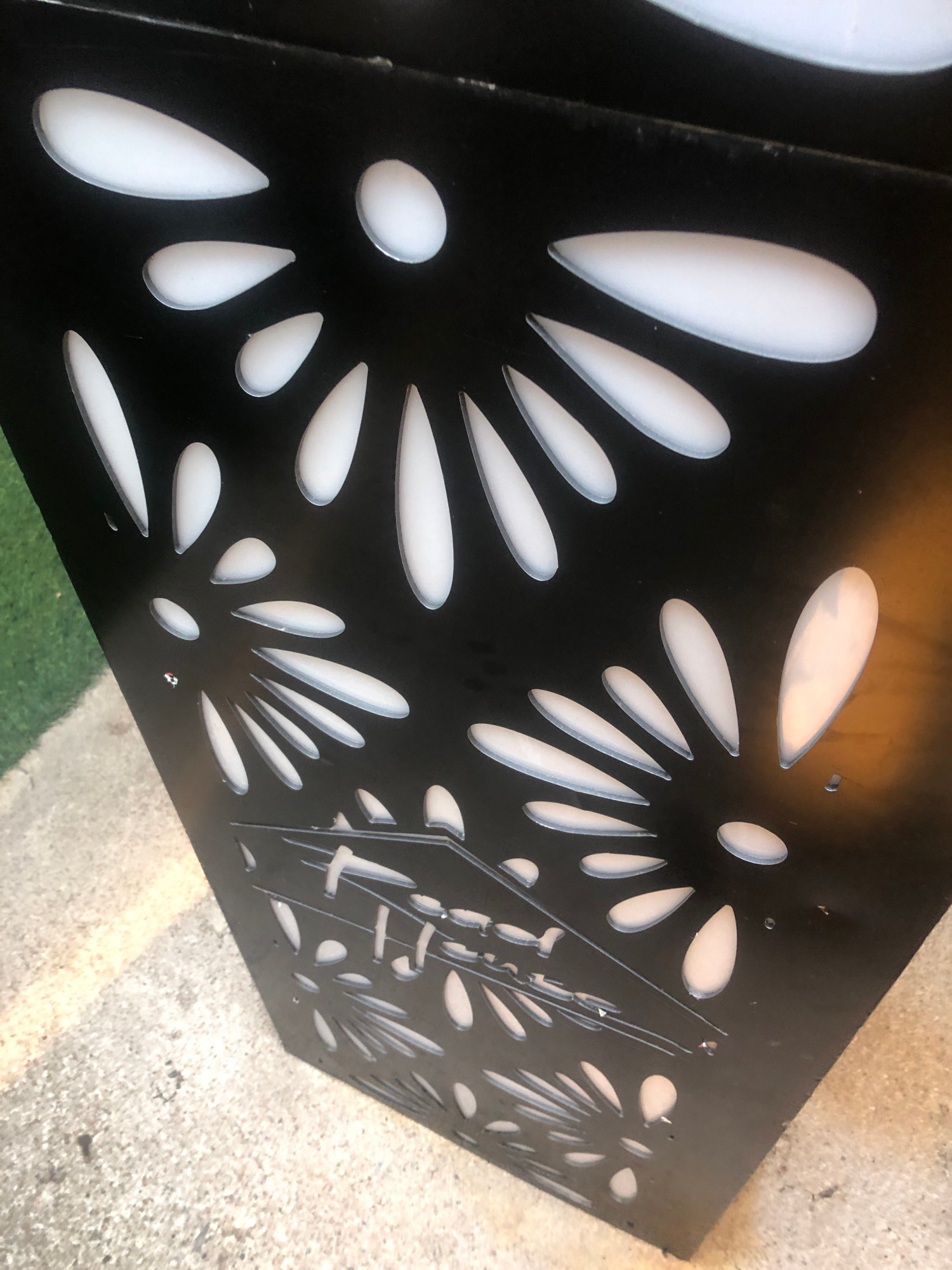 The image shows a close-up of a black, angular object with a decorative pattern of white, elongated oval shapes cut into its surface. These cutouts appear to be illuminated from behind, creating a contrast between the black material and the white light. The object is positioned outdoors, with a textured, light-colored concrete surface visible at the bottom and a green, grassy area to the left. The lighting suggests it is daytime. The object is likely a planter or a decorative panel. The location is specified as Howayi, Nigeria.