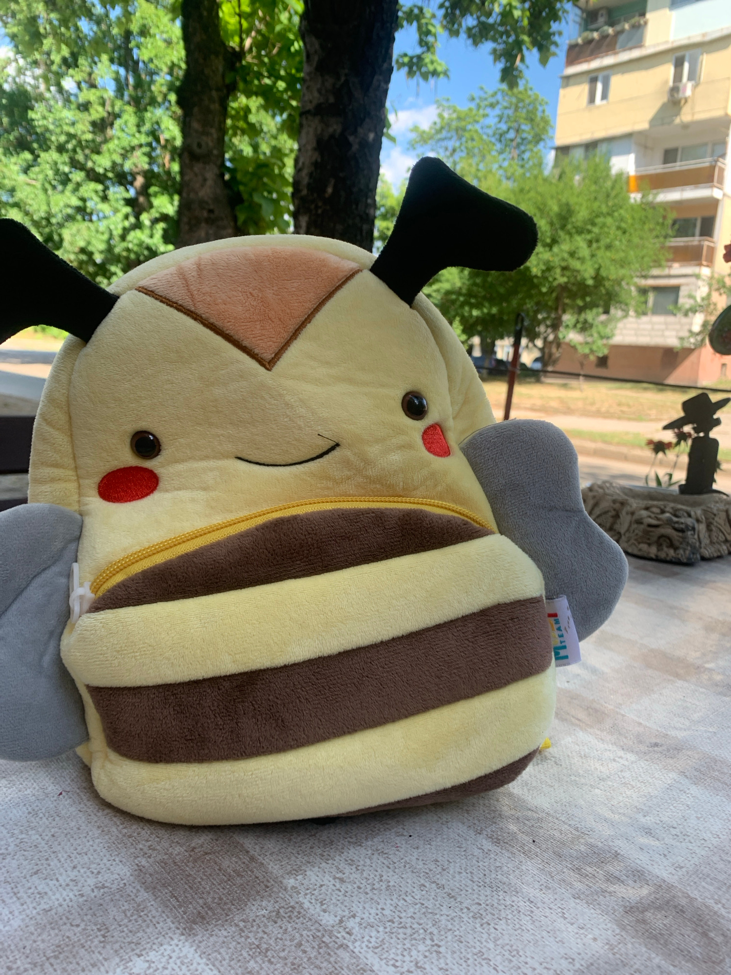 A stuffed bee backpack is placed on a striped tablecloth outdoors, with a blurred background of trees, a building, and a statue. The stuffed toy is yellow and brown, with gray wings and black antennae. A small tag is attached to its side. In the background, a multi-story building with balconies and windows is visible. A silhouetted statue of a person wearing a hat is positioned near a decorative stone structure. The scene appears to be in Vidin, Bulgaria, given the characteristic architecture of the building and the overall setting. The lighting suggests it is daytime.
