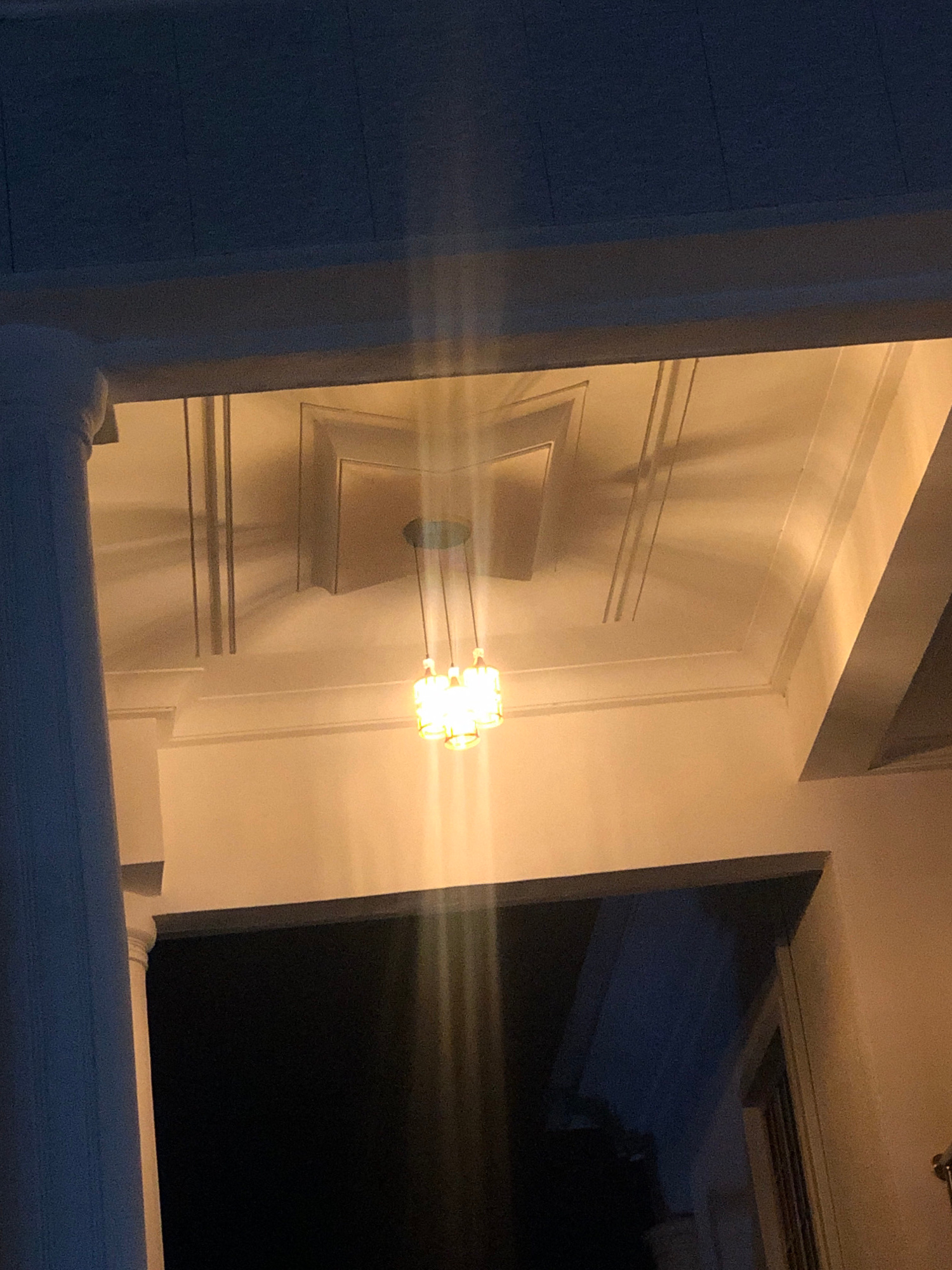 The image captures an interior view looking upwards towards a ceiling. A modern chandelier with multiple illuminated bulbs hangs from the center of the ceiling. The ceiling itself features recessed geometric paneling with subtle shadow lines. To the left, a portion of a white, fluted column is visible, suggesting it is part of a porch or entryway. The lighting is warm and casts distinct shadows across the ceiling. The upper portion of the image is dark, indicating it might be nighttime or an area with minimal natural light. There is no visible indication of the image's location in Bar Jirgi Summa, Nigeria, or any people or specific actions taking place.