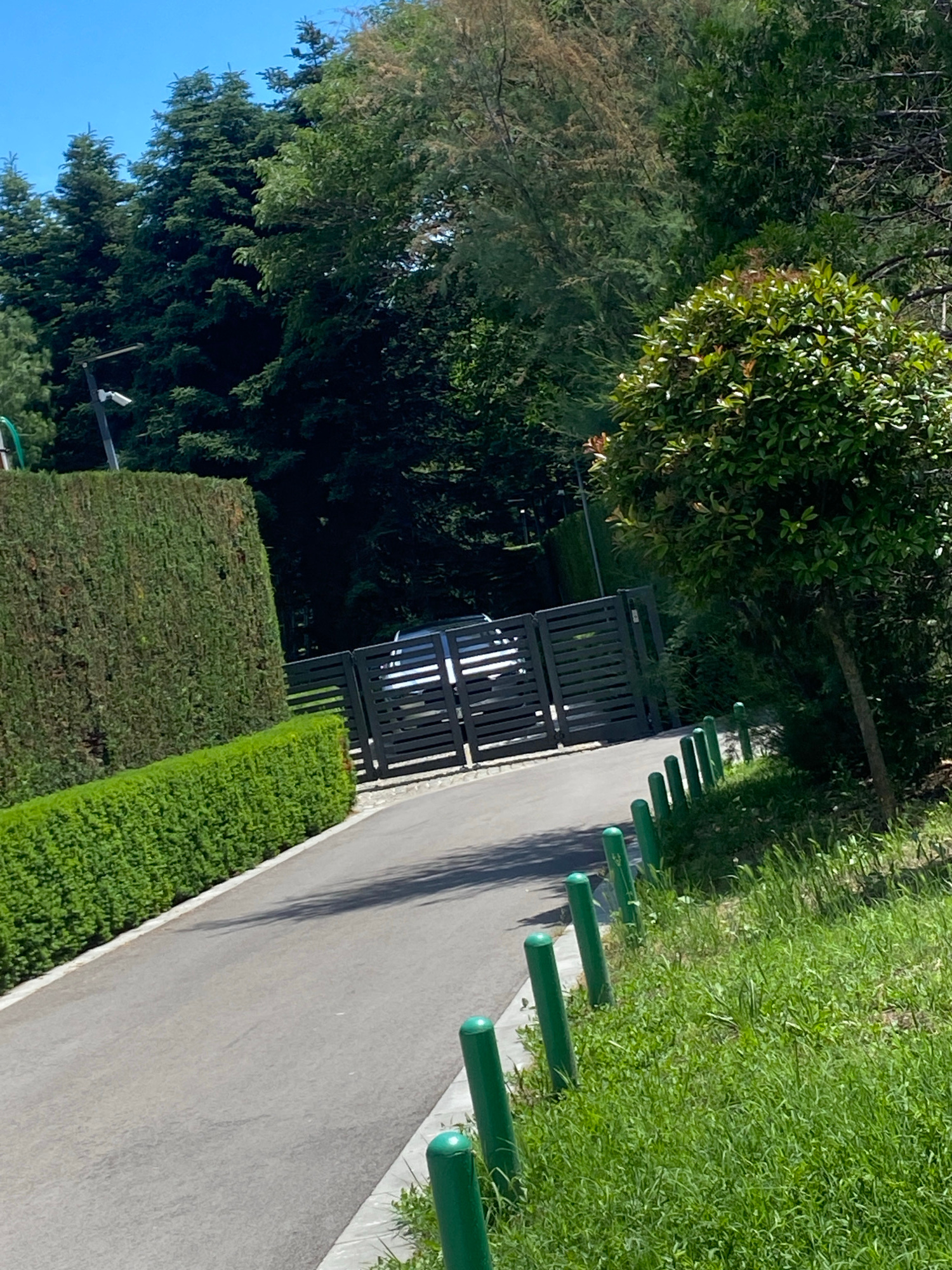 The image shows a paved path with a row of green bollards on one side, separating it from a grassy area. On the other side of the path, there is a black metal fence. Beyond the fence, there are trees and bushes, some of which appear to be manicured hedges. A white vehicle is partially visible through the fence. The scene appears to be outdoors on a sunny day, with shadows cast on the path, suggesting it is daytime. The overall setting suggests a park, a residential area, or a public space with landscaping. No people are visible in the image. There is no visible text. The context provided indicates the location is Varna, Bulgaria.