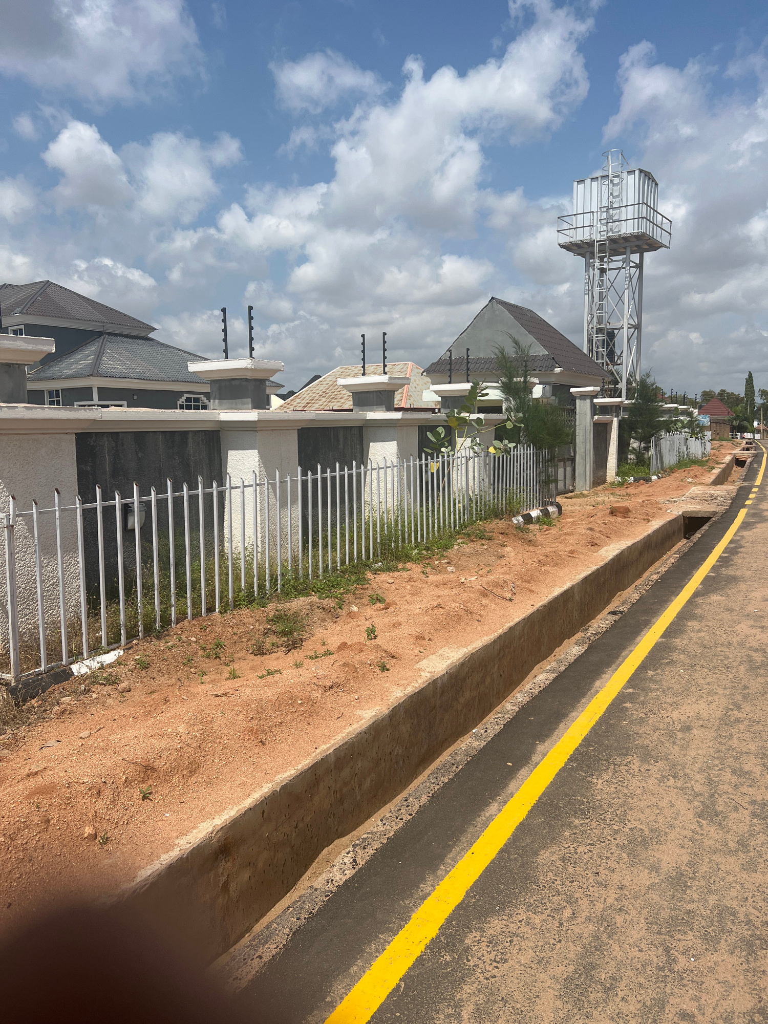 This image depicts a street scene in Howayi, Nigeria, likely taken during the day. The orientation of the image is sideways. The foreground features a paved road with a prominent yellow line painted along its edge, separating it from a concrete drainage channel. The channel is filled with reddish-brown soil and sparse vegetation. Beyond the channel, a white metal fence encloses a property.
Within the property, several modern houses are visible, characterized by their dark-tiled roofs and light-colored textured walls. Some houses have windows with white frames. Decorative elements such as short pillars topped with dark spheres are present along the fence line.
Dominating the left side of the image, a tall metal water tower stands against a bright blue sky dotted with white clouds. The tower has a white tank at its top, supported by a complex metal framework. Some greenery, possibly trees or large bushes, can be seen peeking over the fence and between the houses.
The overall impression is one of a developing residential area with modern infrastructure. The weather appears fair, with ample sunlight casting shadows, suggesting it is likely daytime. There are no people visible in the image. No text is discernible.