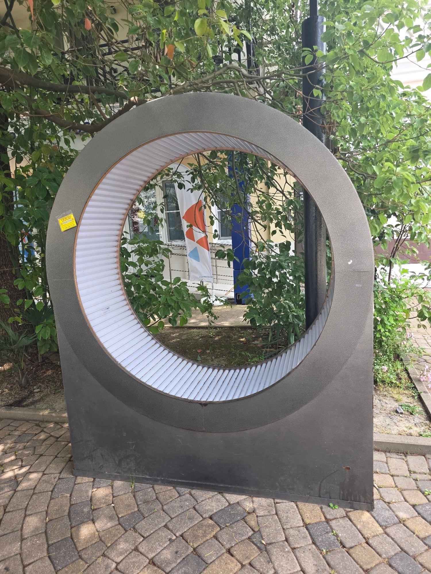 An outdoor circular sculptural element with a ribbed white interior is positioned on a patterned stone walkway, surrounded by greenery. The sculpture is dark gray and appears to be made of metal or a similar composite material. A yellow sticker is affixed to the left side of the sculpture. Through the circular opening, a building with windows and a flag featuring orange and blue geometric shapes is visible. The environment suggests a public space within a city, possibly a park or courtyard. The provided information indicates the location is the city of Sochi, Russia.