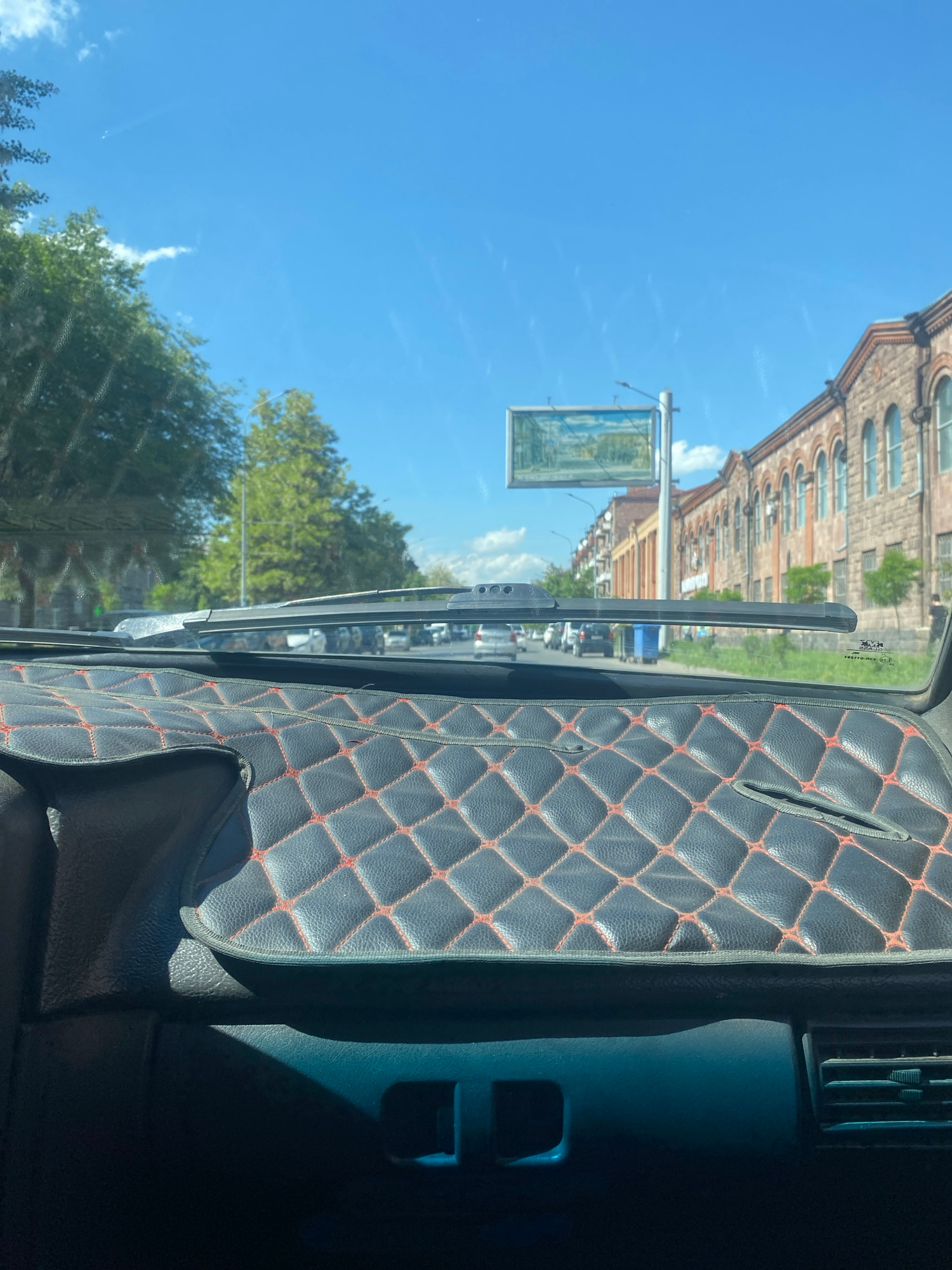 The image shows a view from inside a car, looking forward through the windshield. The dashboard is partially visible on the right side, covered with a diamond-stitched black material with red trim. A windshield wiper is positioned on the left side of the windshield.
Outside the car, the scene is a sunny day with a clear blue sky. A multi-story brick building with arched windows is visible in the background on the left. A signpost with a rectangular sign featuring a picture is also present in front of the building. Green trees are scattered throughout the scene, and some clouds are visible in the sky. The overall impression is of a bright, clear day in an urban or semi-urban environment. There is no visible activity or event taking place, other than the implied movement of the car. The image is oriented vertically, with the sky at the top and the dashboard at the bottom.
