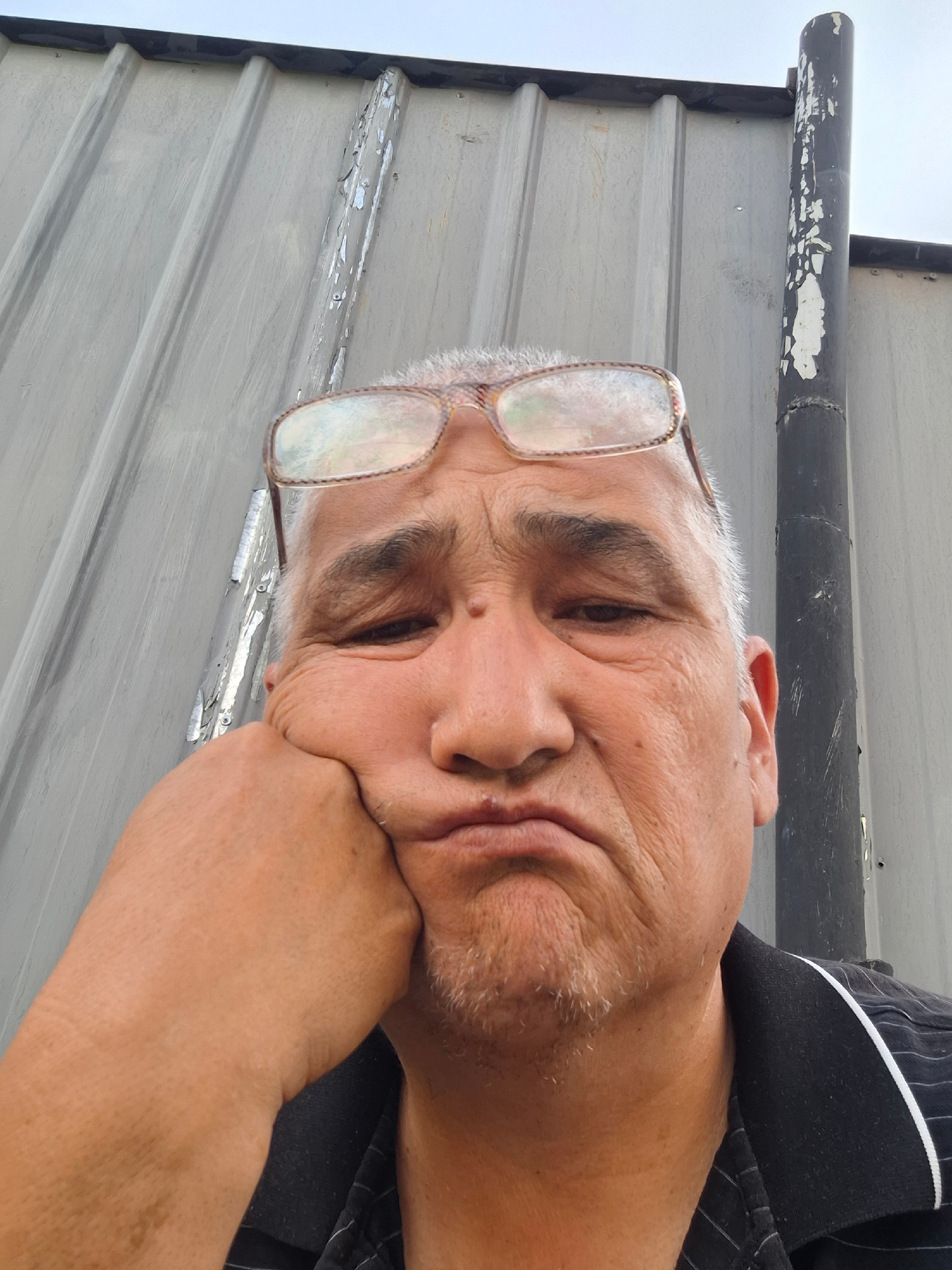 The image is a selfie of an individual with glasses, resting their head on their hand, with a pouty expression. The background appears to be a corrugated metal wall, possibly part of an industrial or storage facility. There is a dark vertical pipe or pole to the right of the individual. The lighting suggests it is daytime, with an overcast sky. The individual is wearing a dark collared shirt with thin white stripes. The image does not contain any recognizable landmarks or elements that would indicate its specific location within the city of Almaty, Kazakhstan.